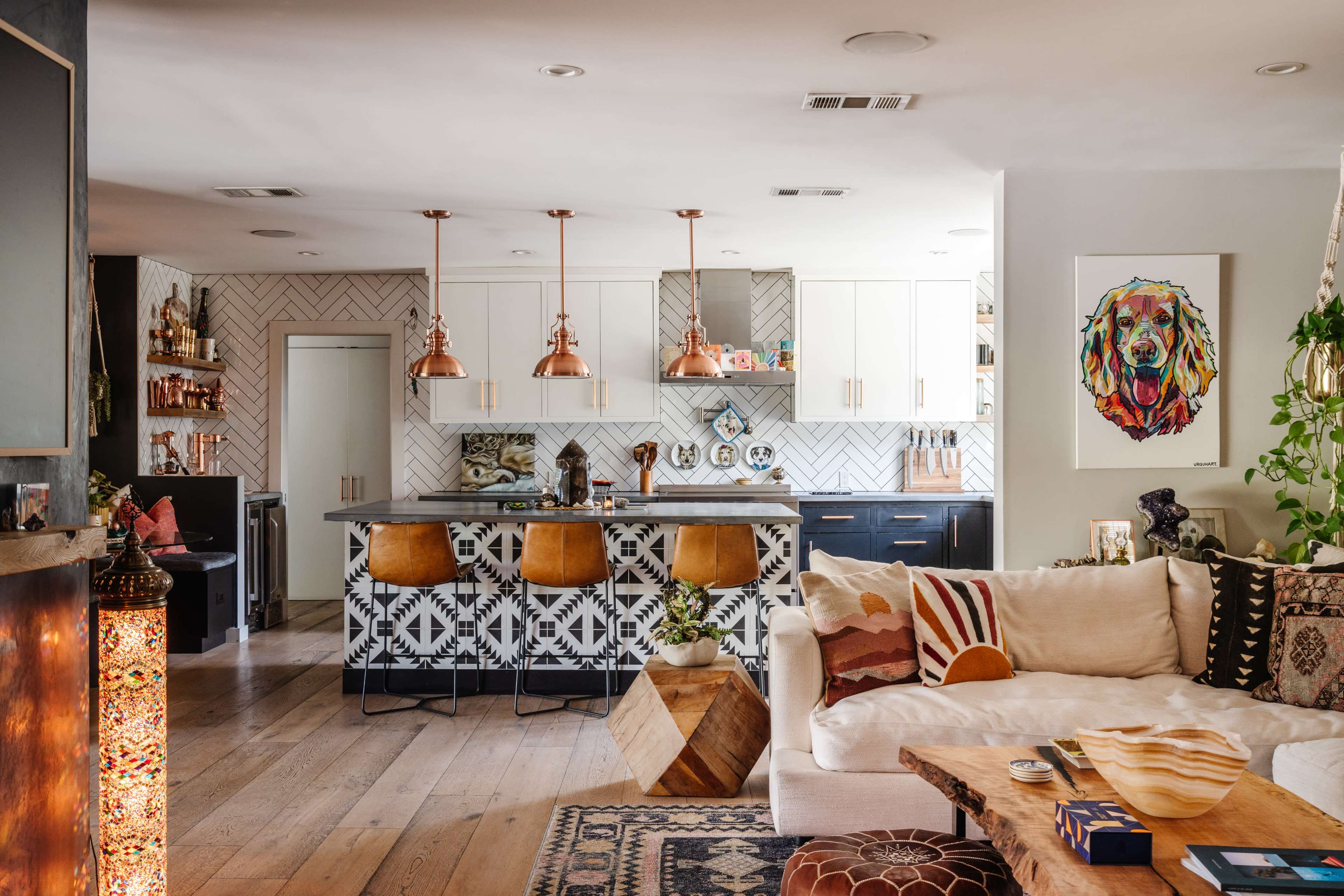 A modern kitchen and living area featuring a kitchen island with copper pendant lights, patterned tiles, and a cozy seating area with a coffee table and decorative pillows.