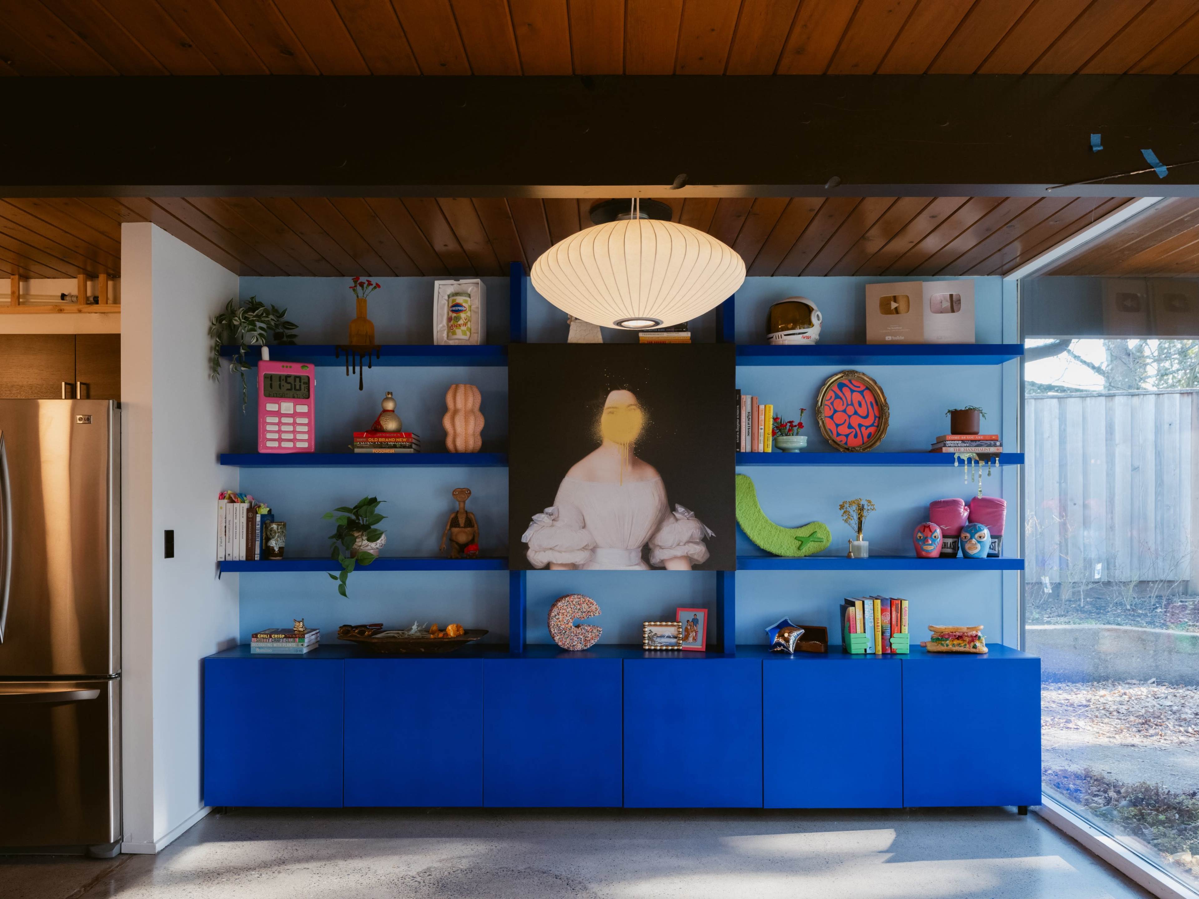 The image depicts a modern blue shelving unit filled with books, decorative items, and a large artwork featuring a figure with a yellow face.