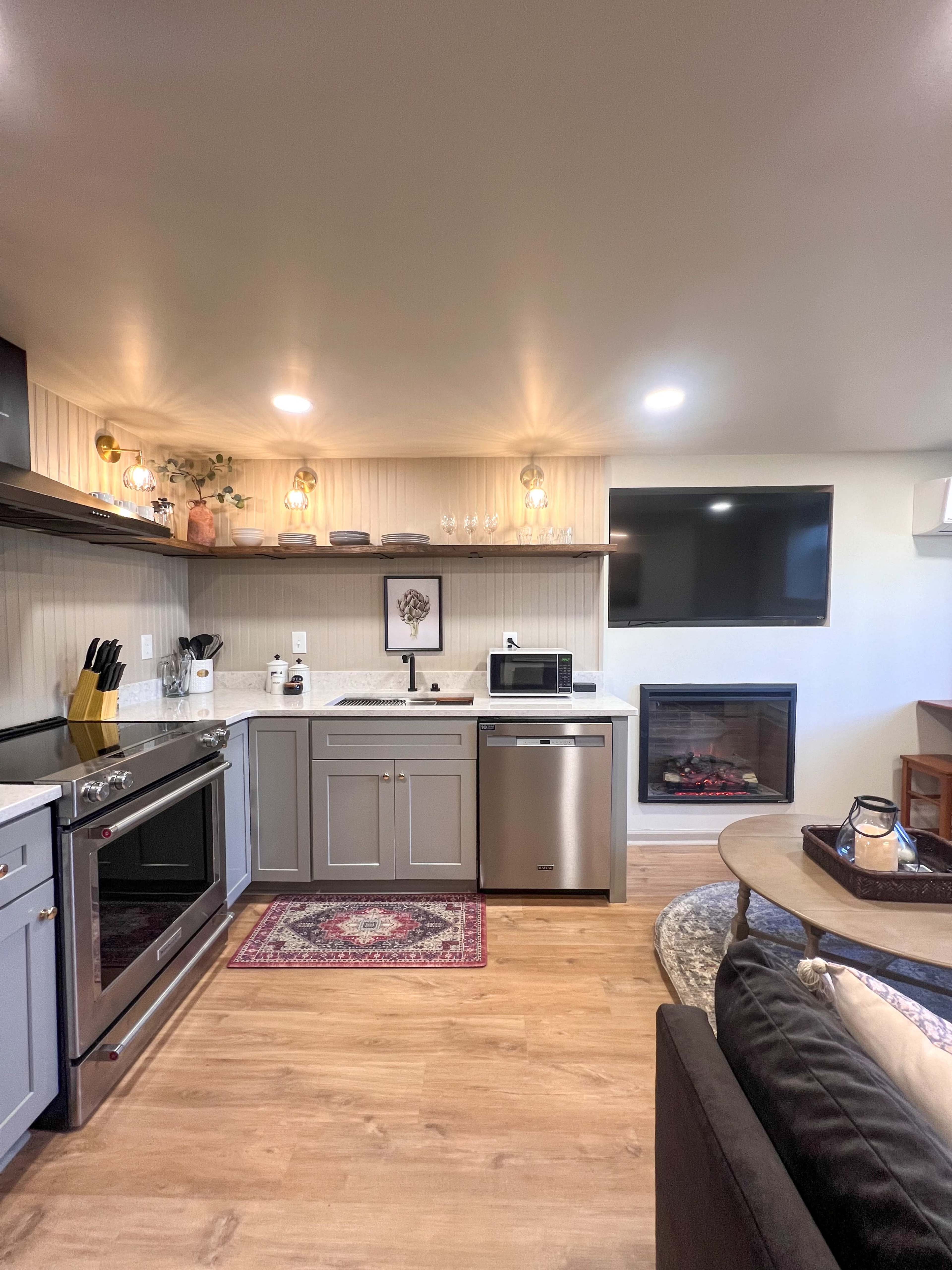 A modern kitchen area with gray cabinets, stainless steel appliances, and a cozy living space featuring a fireplace and a television.