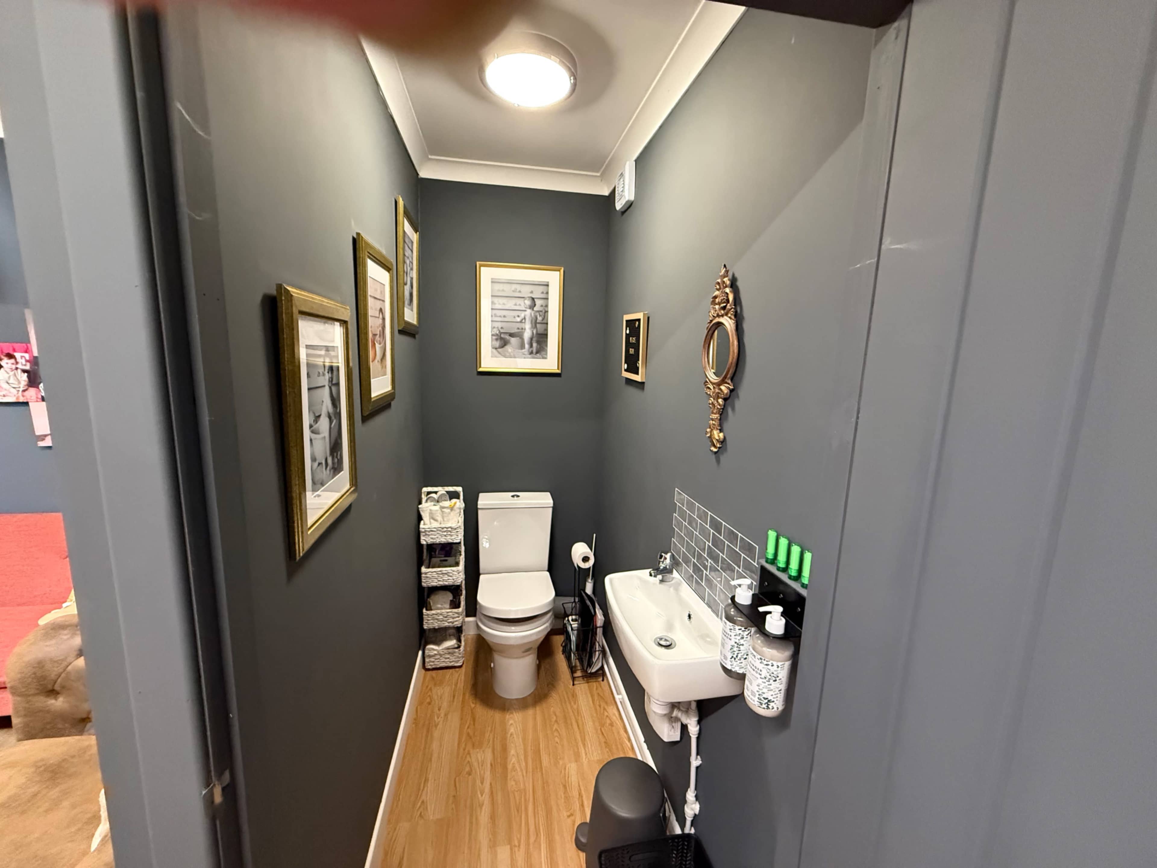 The image shows a small, narrow bathroom featuring a toilet, a sink, and framed pictures on the walls.