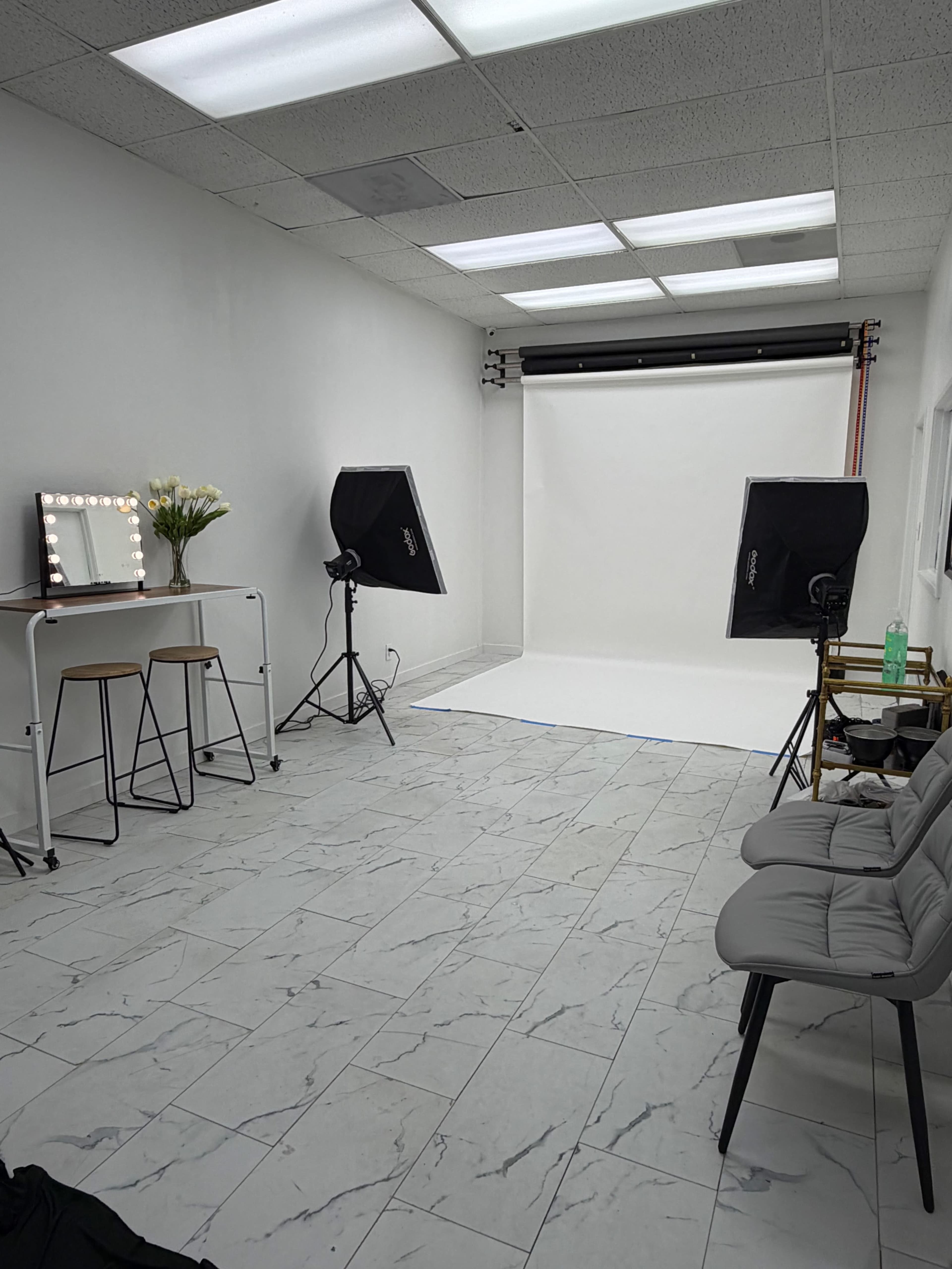 The image shows a minimalistic photography studio with a white backdrop, two black studio lights, a mirror with bulbs, a small table with two stools, and a plant arrangement.