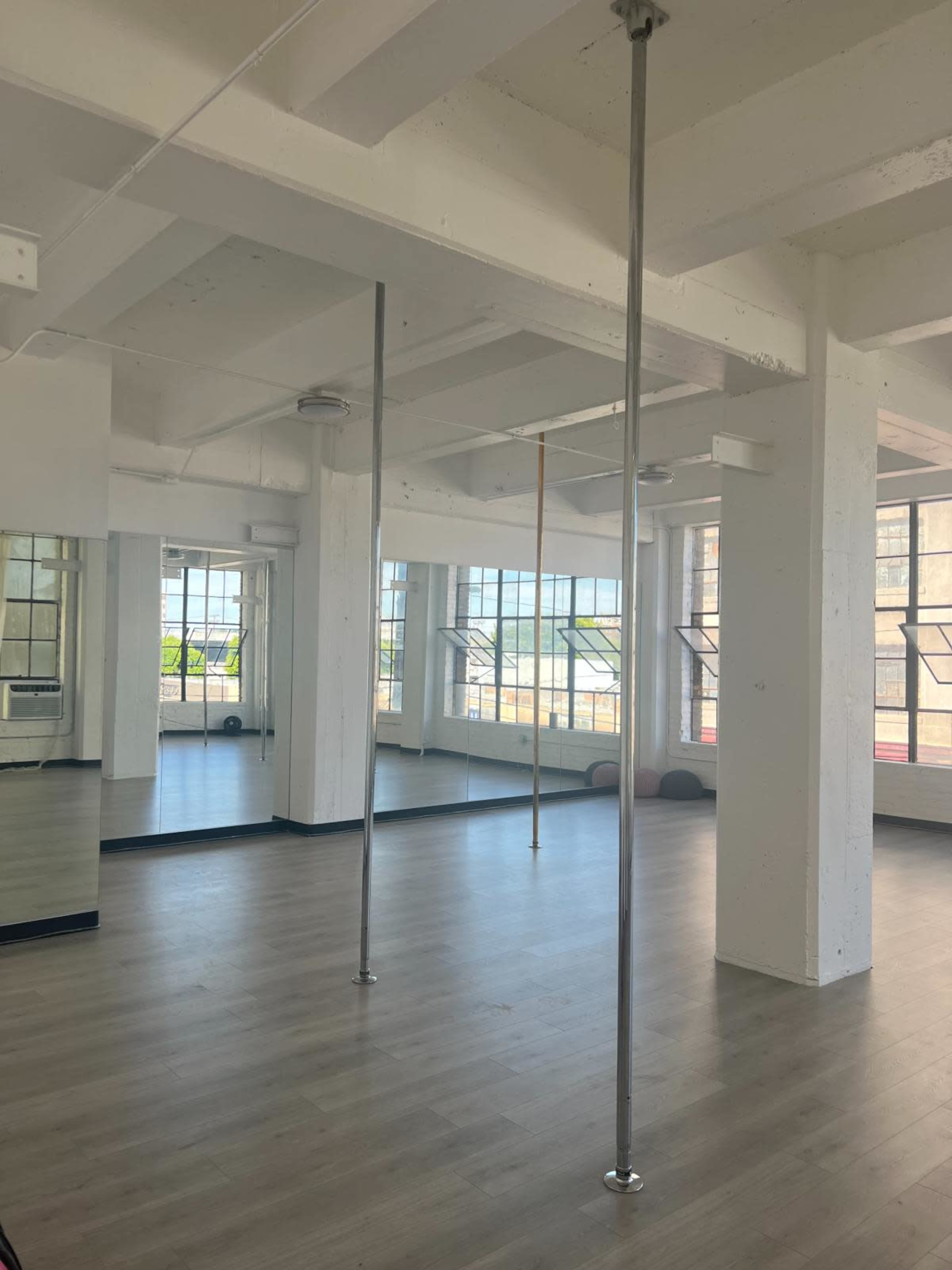 The image shows an empty fitness studio with several vertical poles and large windows allowing natural light to enter.
