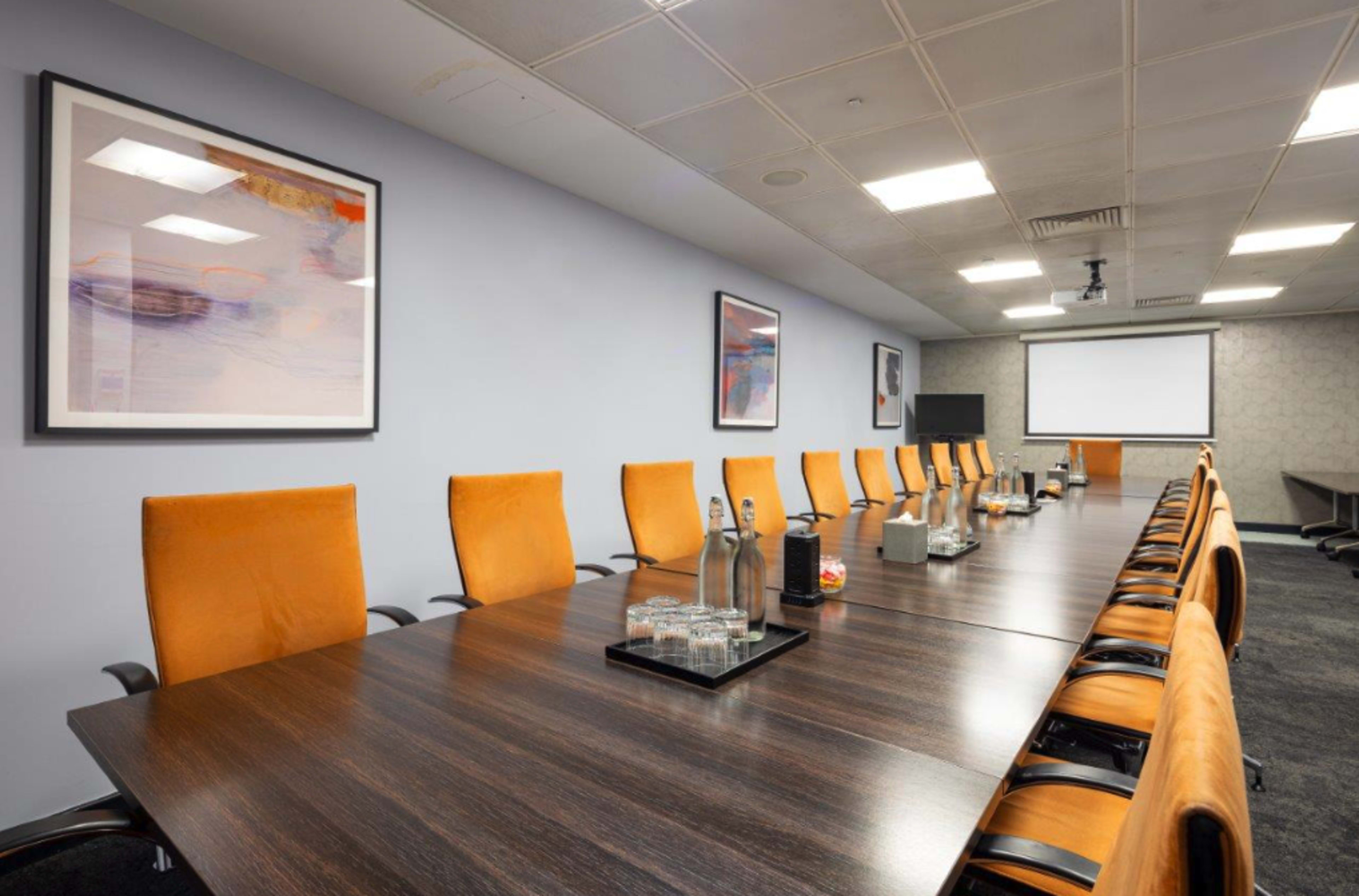 A large conference room features a long table surrounded by orange chairs, with several bottles of water and snacks on the table, and a projector screen at the front.