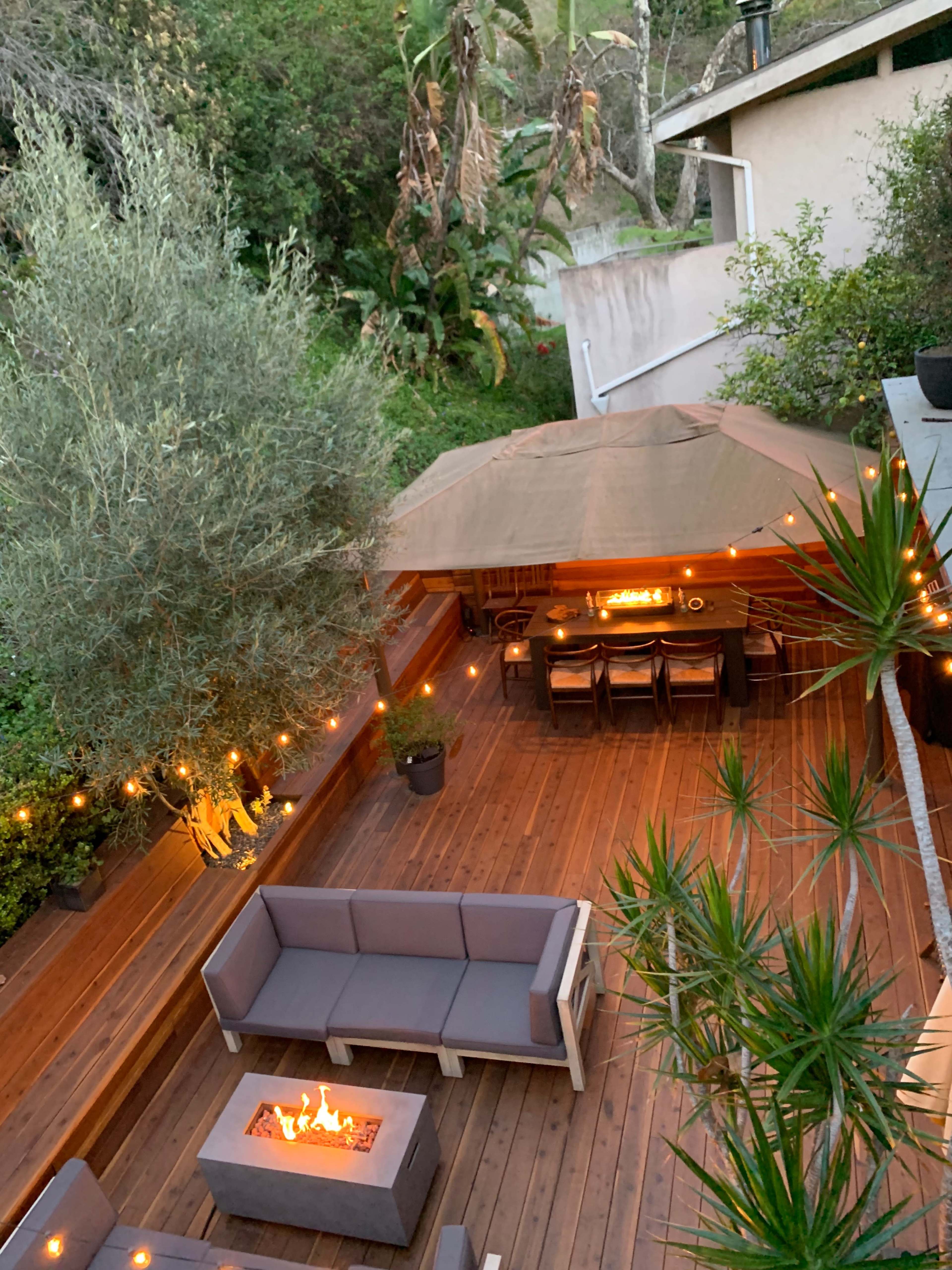 A wooden deck features a fire pit surrounded by seating and a canopy-covered bar area, illuminated by string lights.