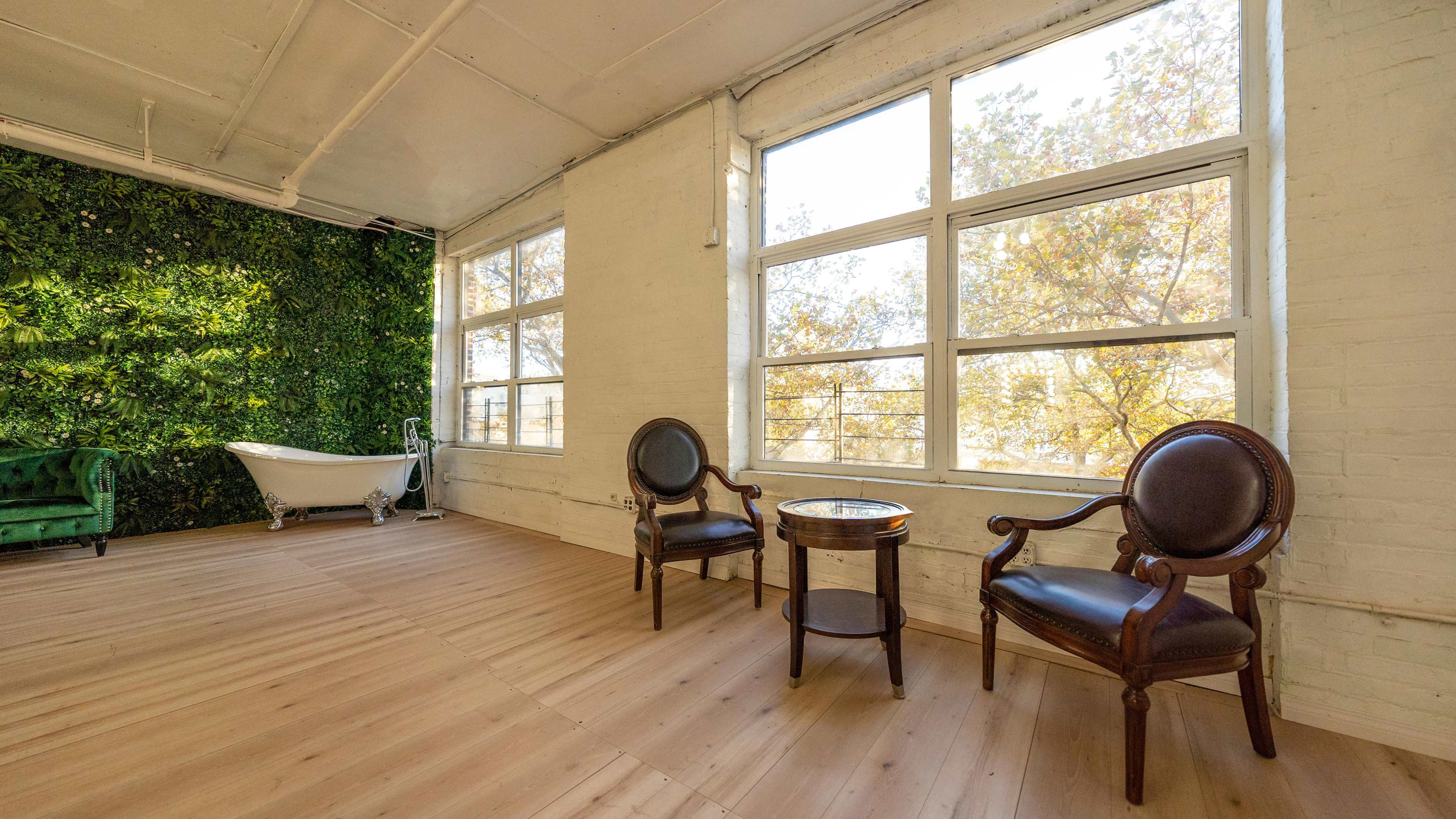The image shows a bright, spacious room with large windows, two vintage chairs, a small table, and a green plant wall in the background.