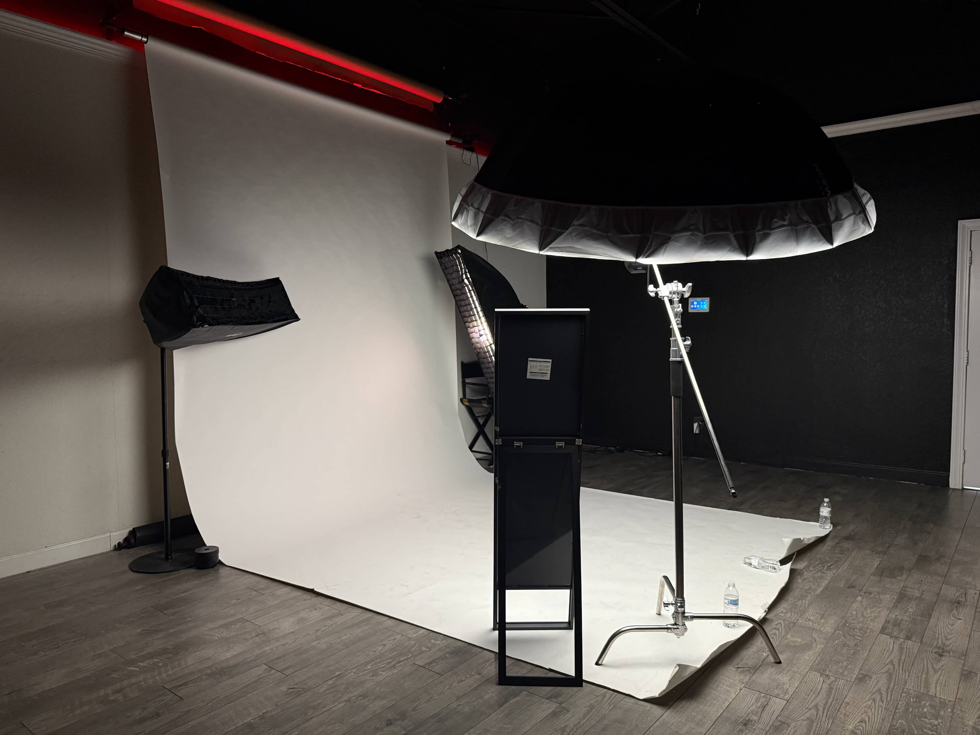 The image shows a photography studio setup with a white backdrop, softbox lighting, and a black monitor on a stand.