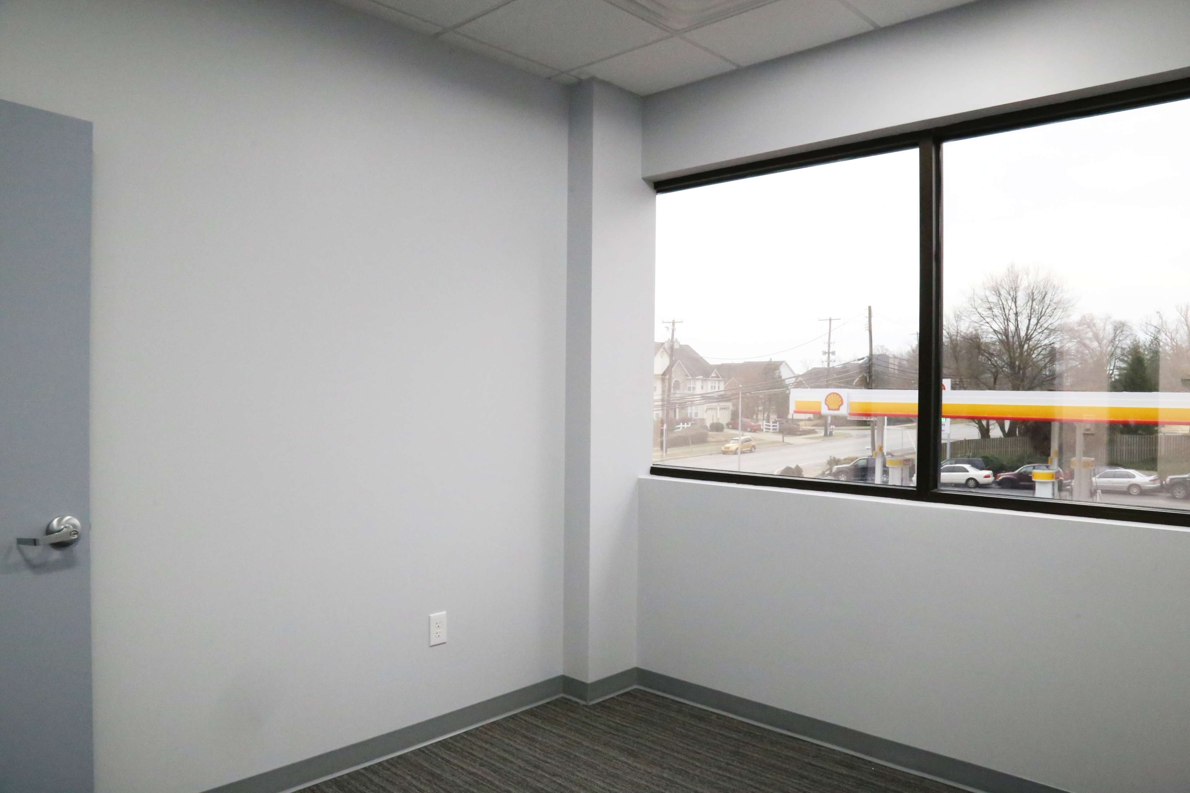 A sparsely furnished office space with a gray wall and a window overlooking a street and a gas station.