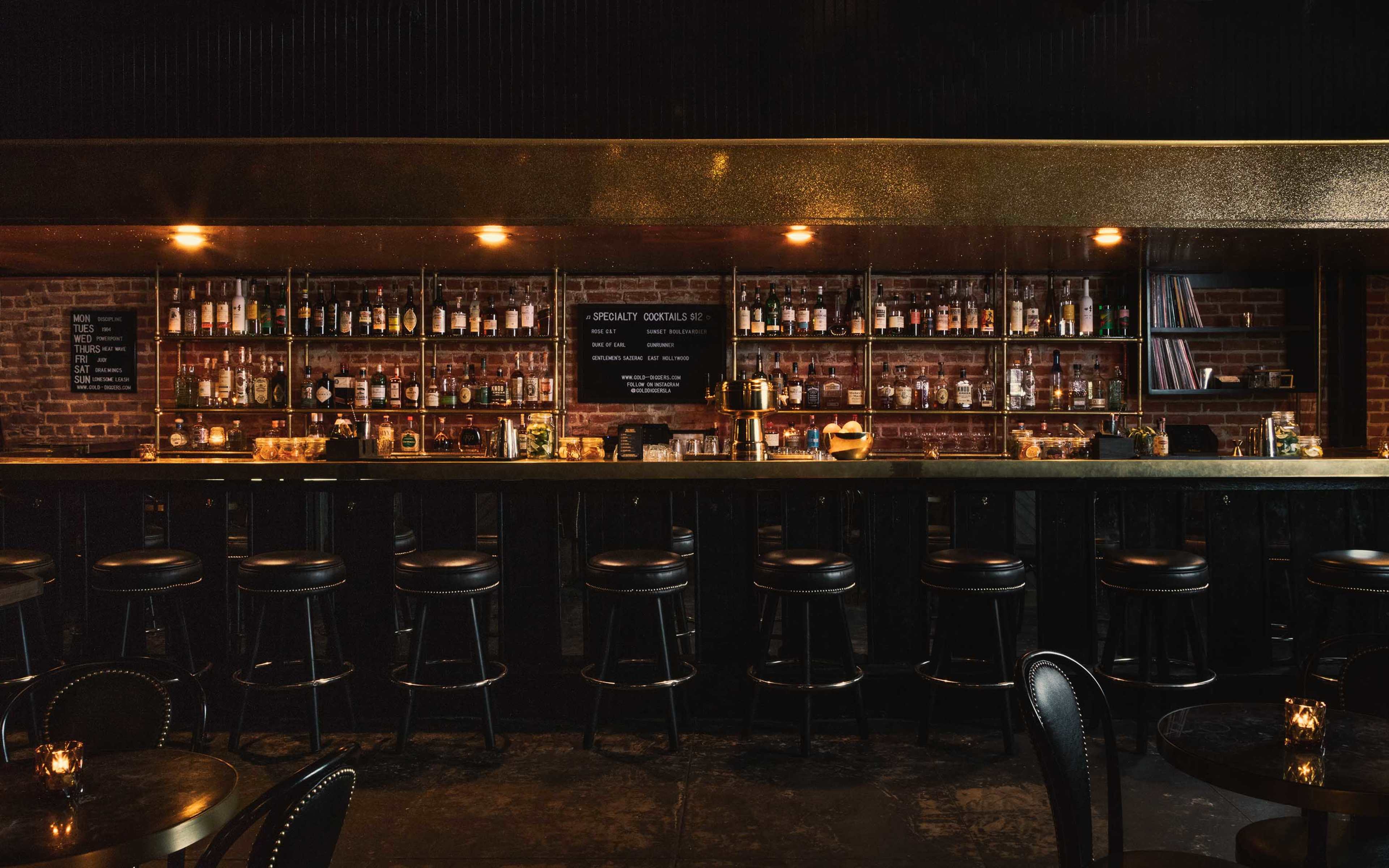 A dimly lit bar with a long counter, featuring shelves filled with various bottles and a few small tables with chairs.