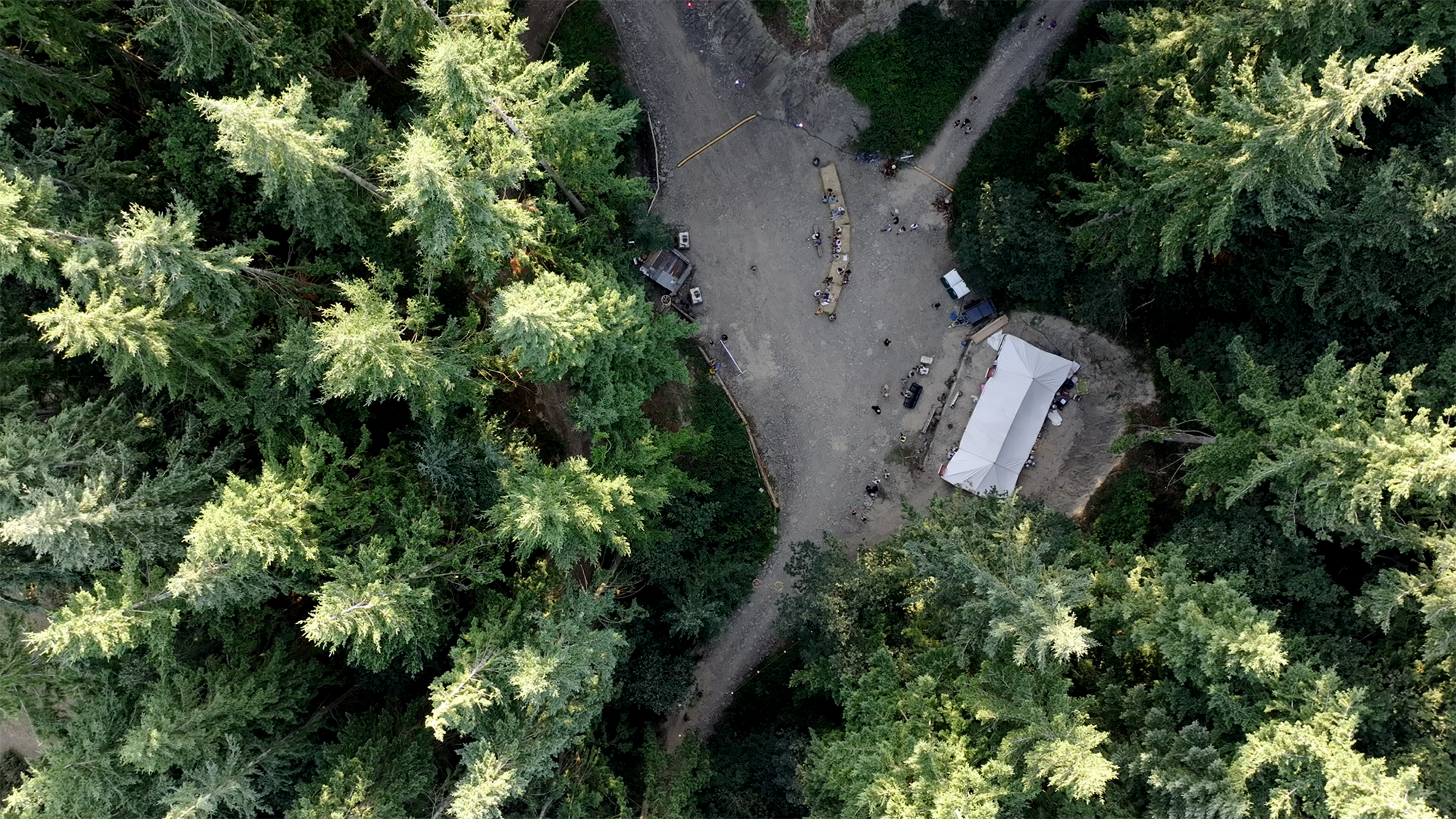 An aerial view shows a clearing surrounded by tall trees, with a white tent and several scattered people and equipment on the ground.