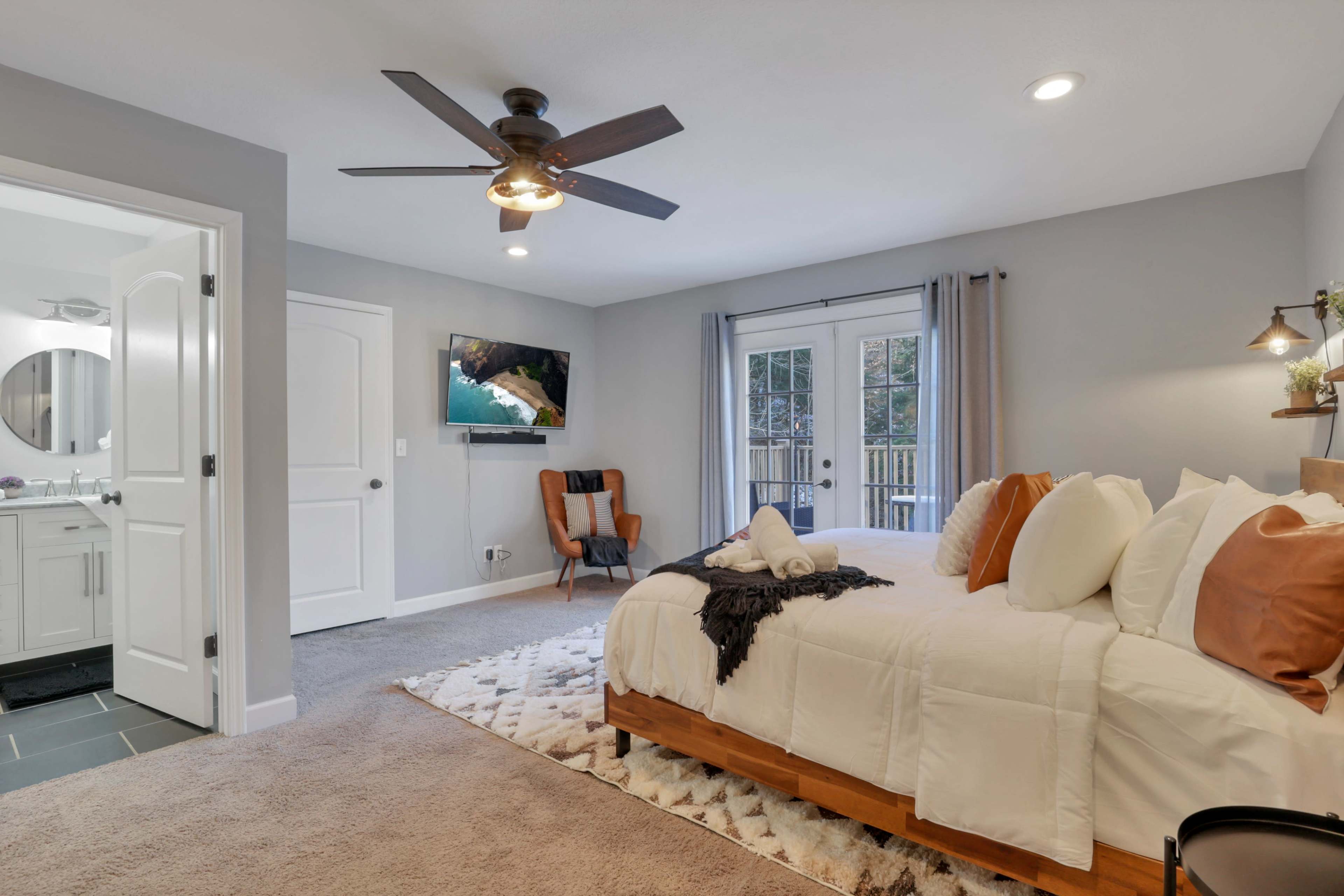 A bedroom with a wooden bed, a flat-screen TV mounted on the wall, a chair in one corner, and an attached bathroom visible through an open door.