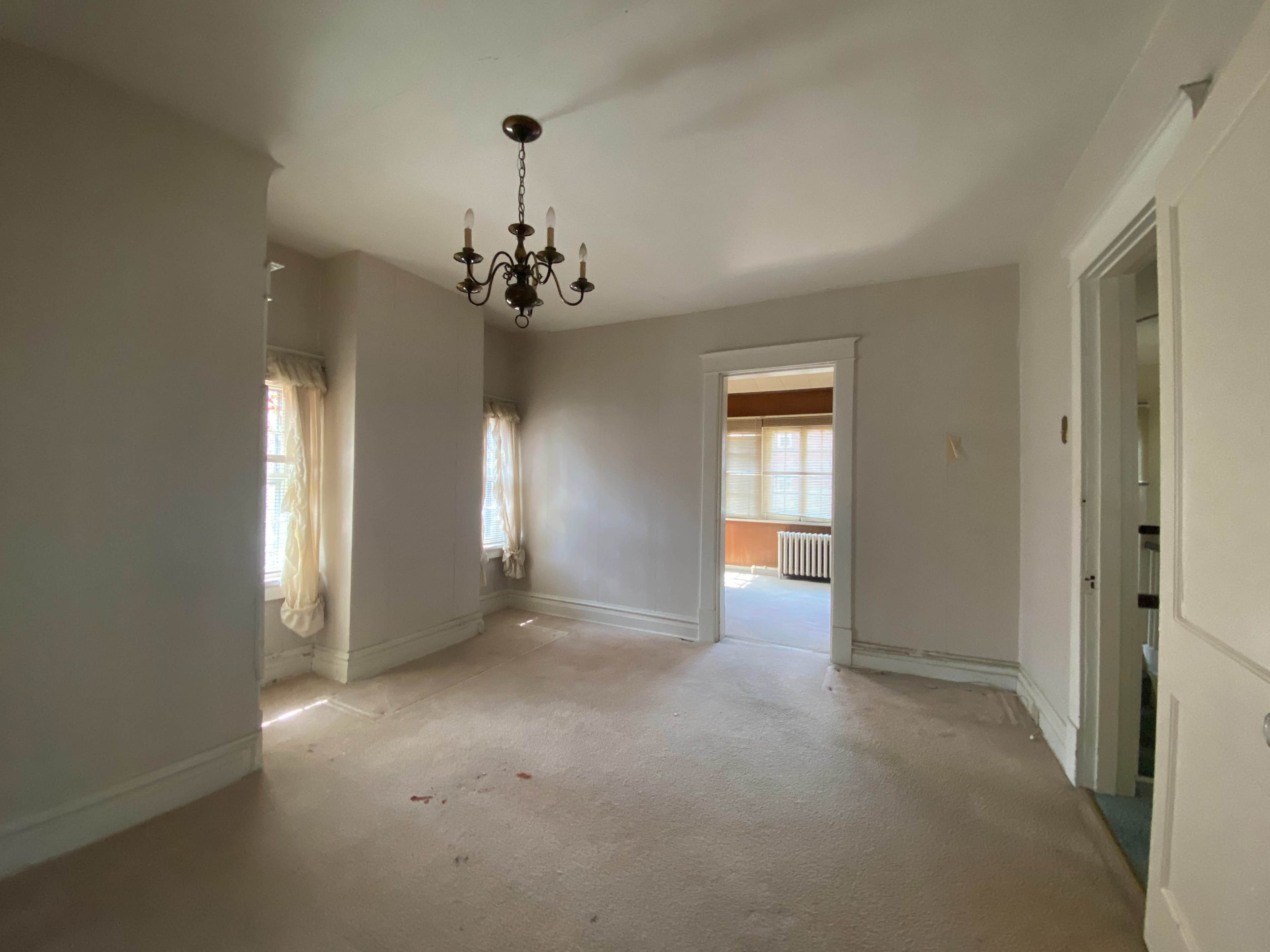 A vacant room features a chandelier, two windows with curtains, and an adjacent room visible through an open doorway.