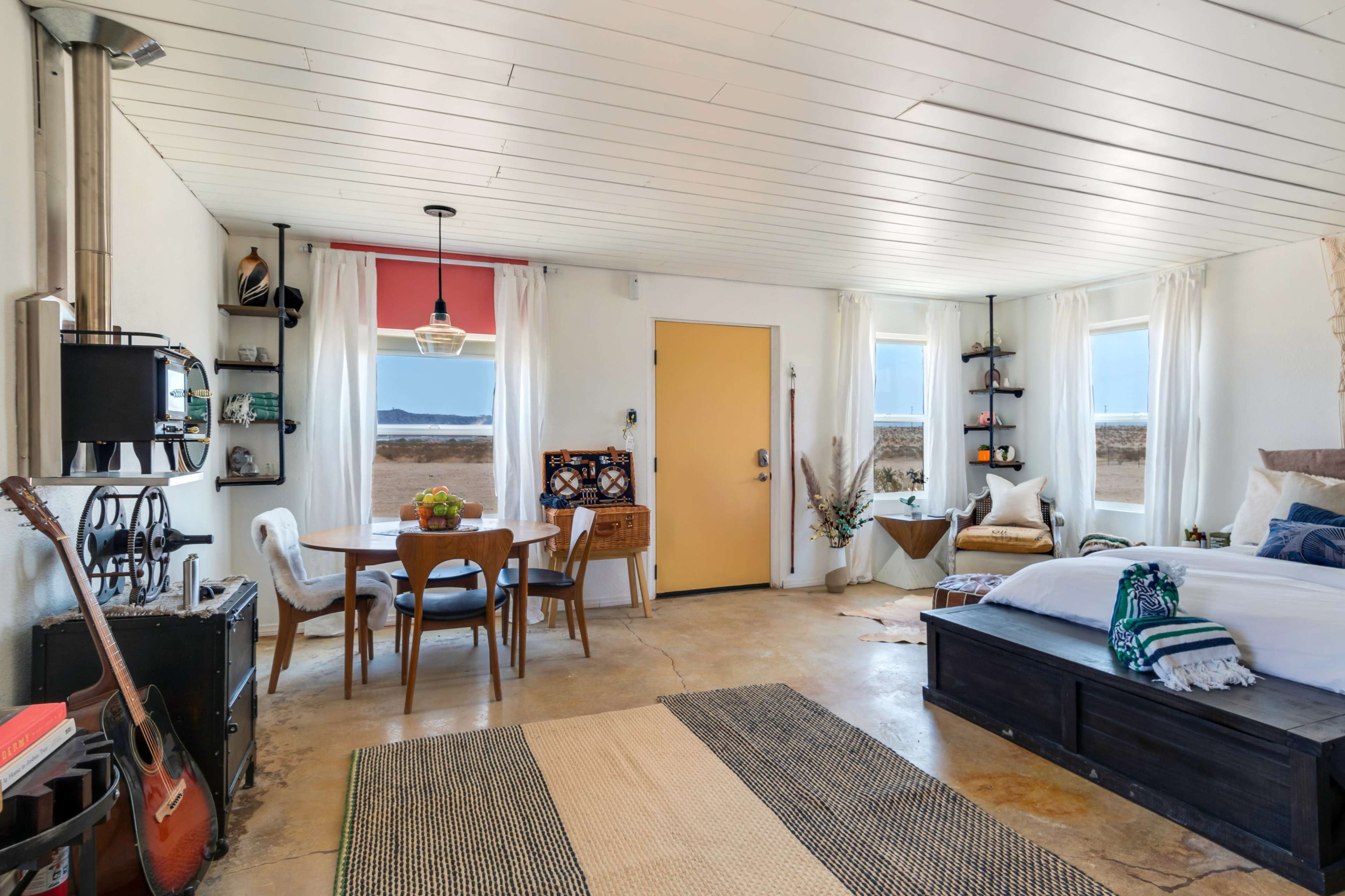 A cozy interior of a small room that includes a bed, dining table, and large windows with a view of the outdoors.