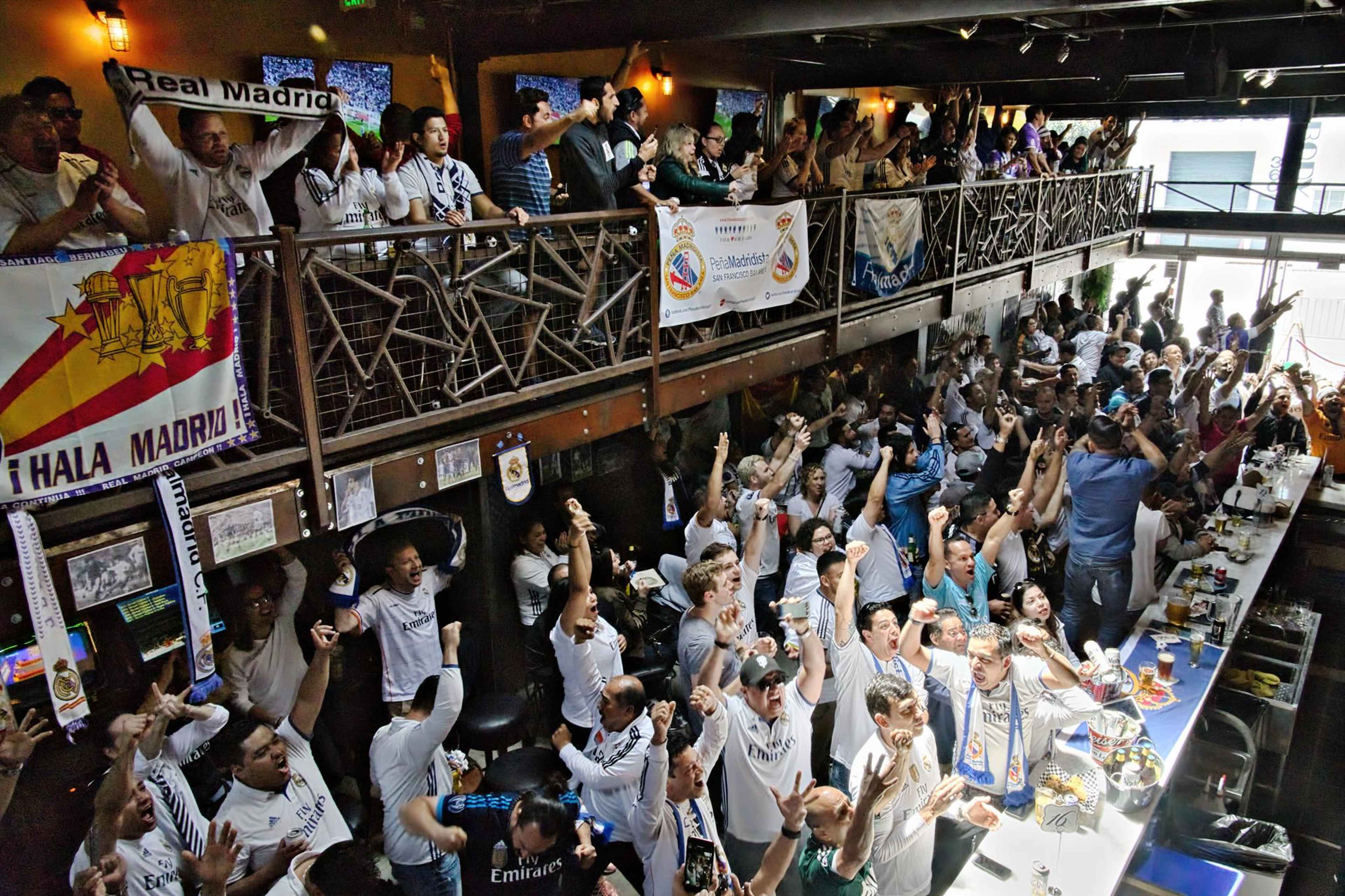 A large crowd of fans wearing white jerseys and holding banners celebrates enthusiastically in a busy sports bar during a soccer match.