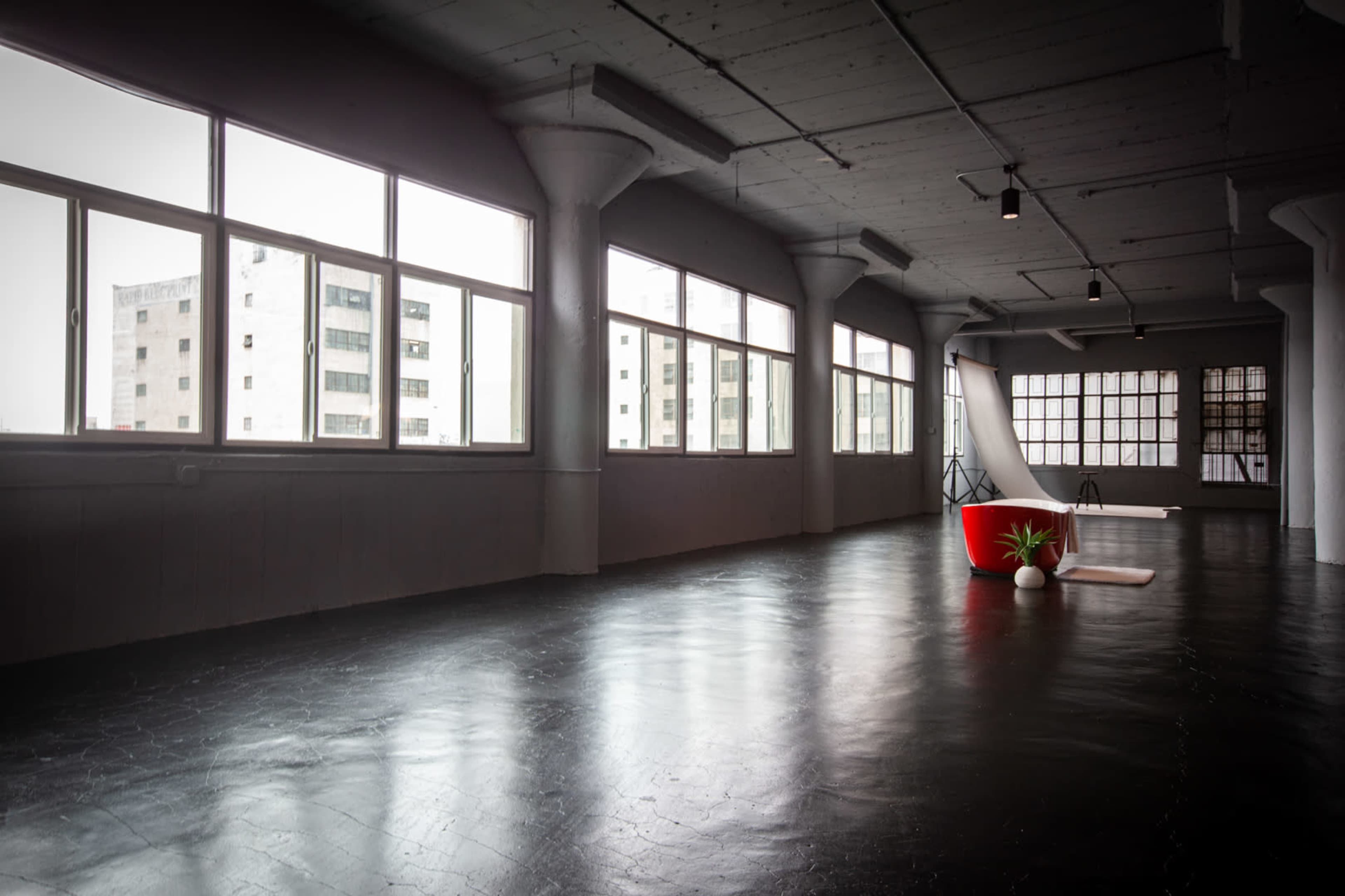 The image shows a spacious, minimalist room with large windows and a polished black floor, featuring a red chair and a small plant.