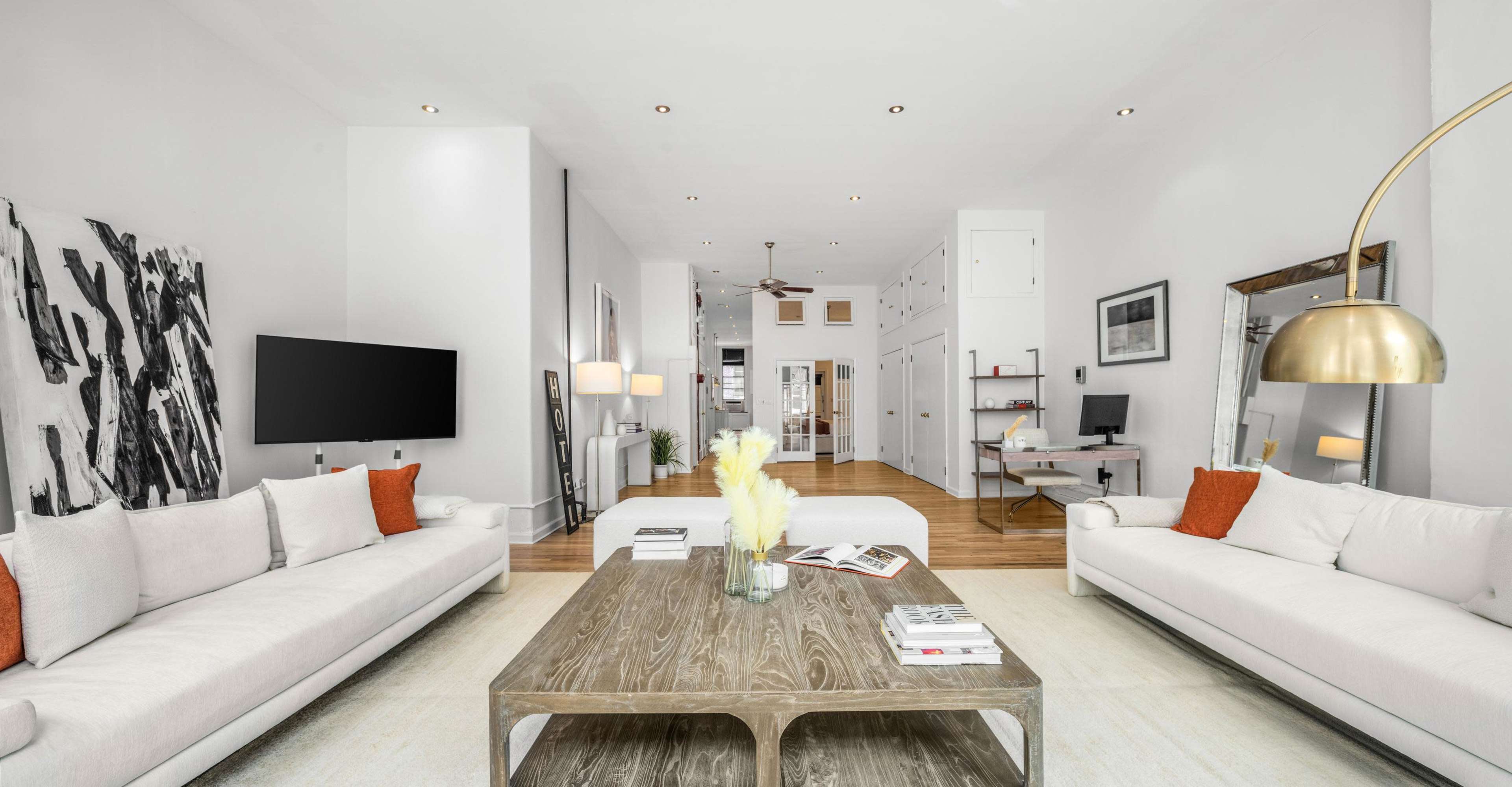 A spacious living room with white sofas, a wooden coffee table, and a wall-mounted television, leading into a bright hallway with additional rooms visible in the background.