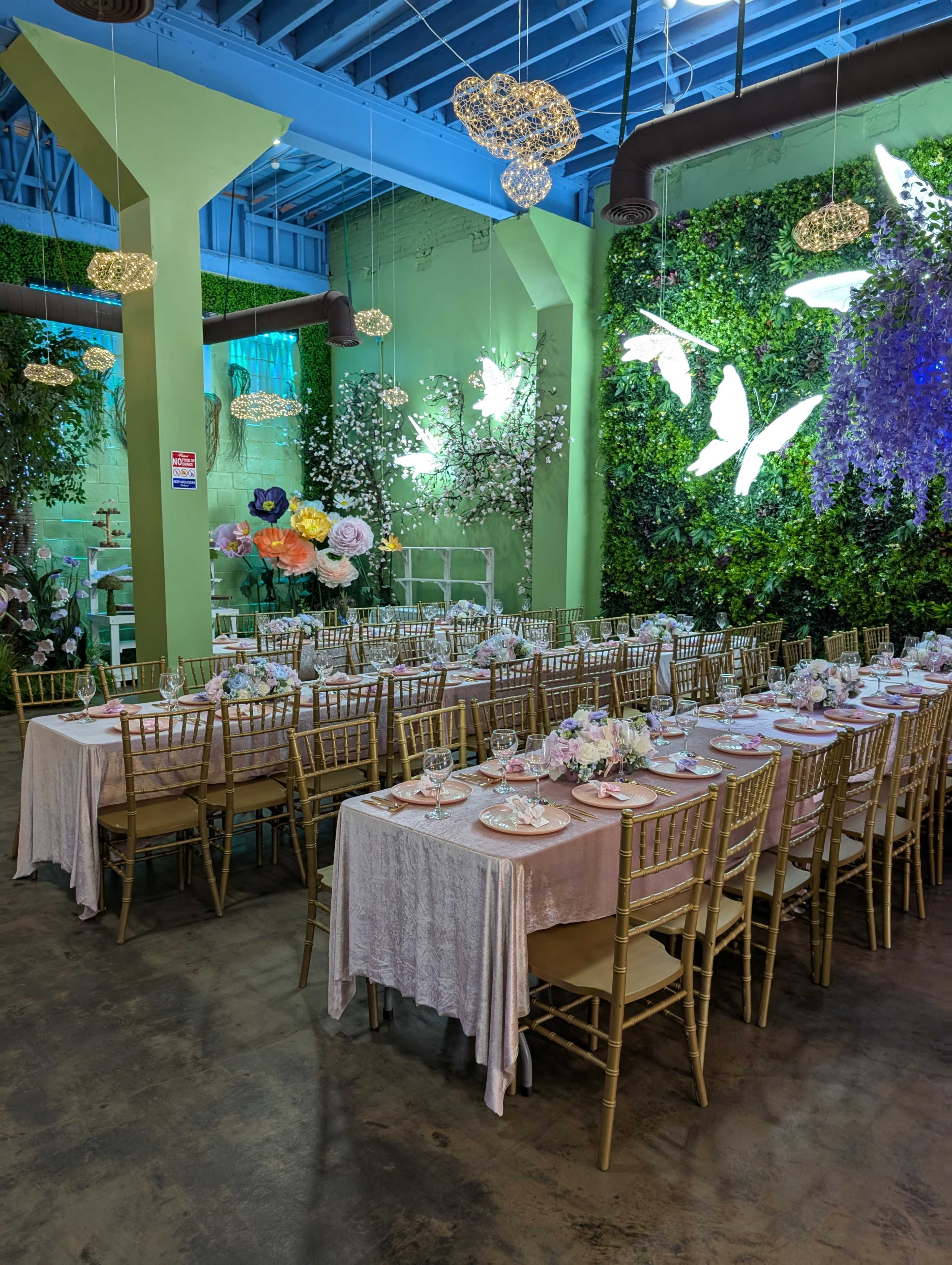 The image depicts a decorated dining area with multiple long tables set with plates and glasses, surrounded by greenery and illuminated by hanging lights.