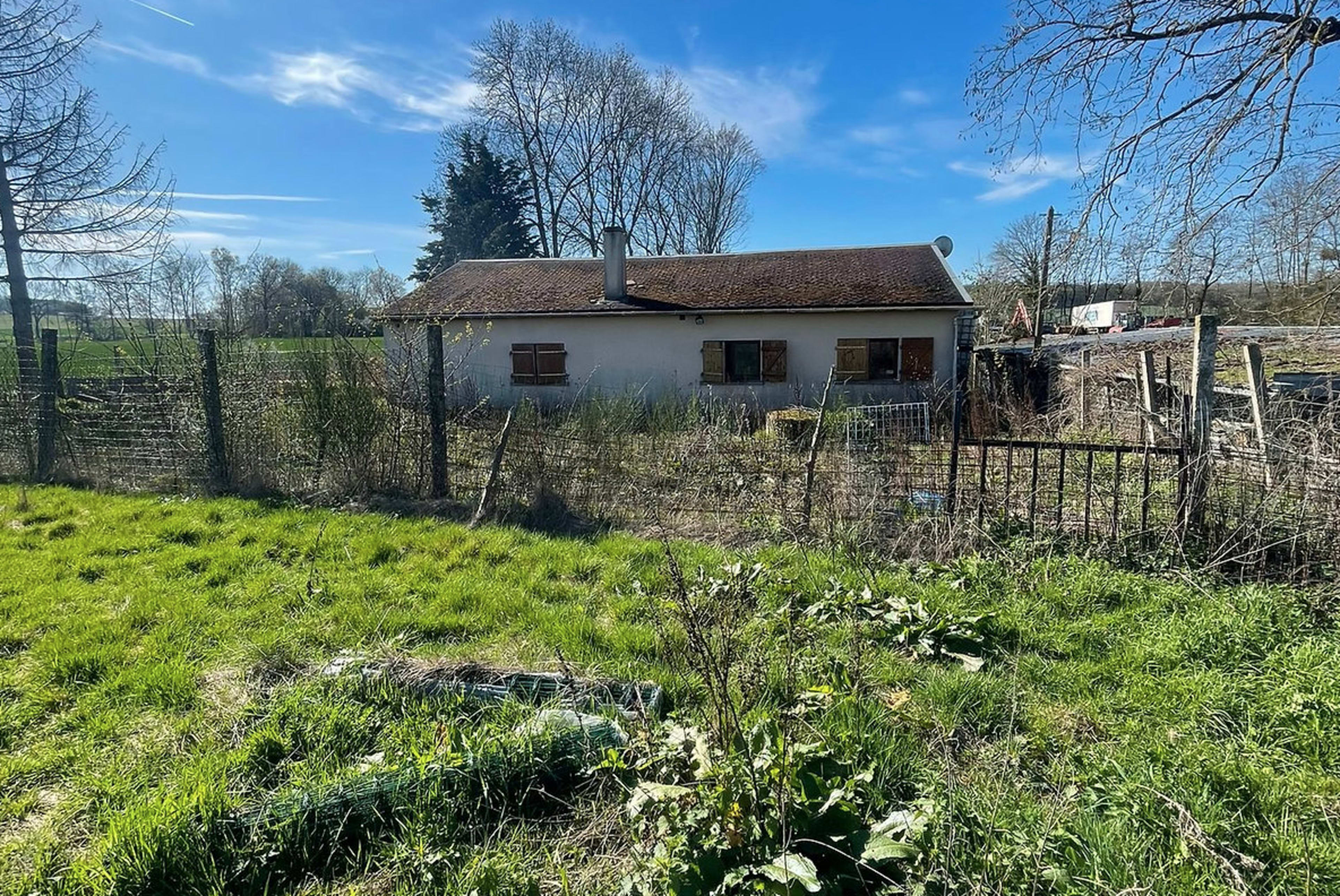 A small, unkempt house is situated in a grassy area surrounded by a fence under a clear blue sky.