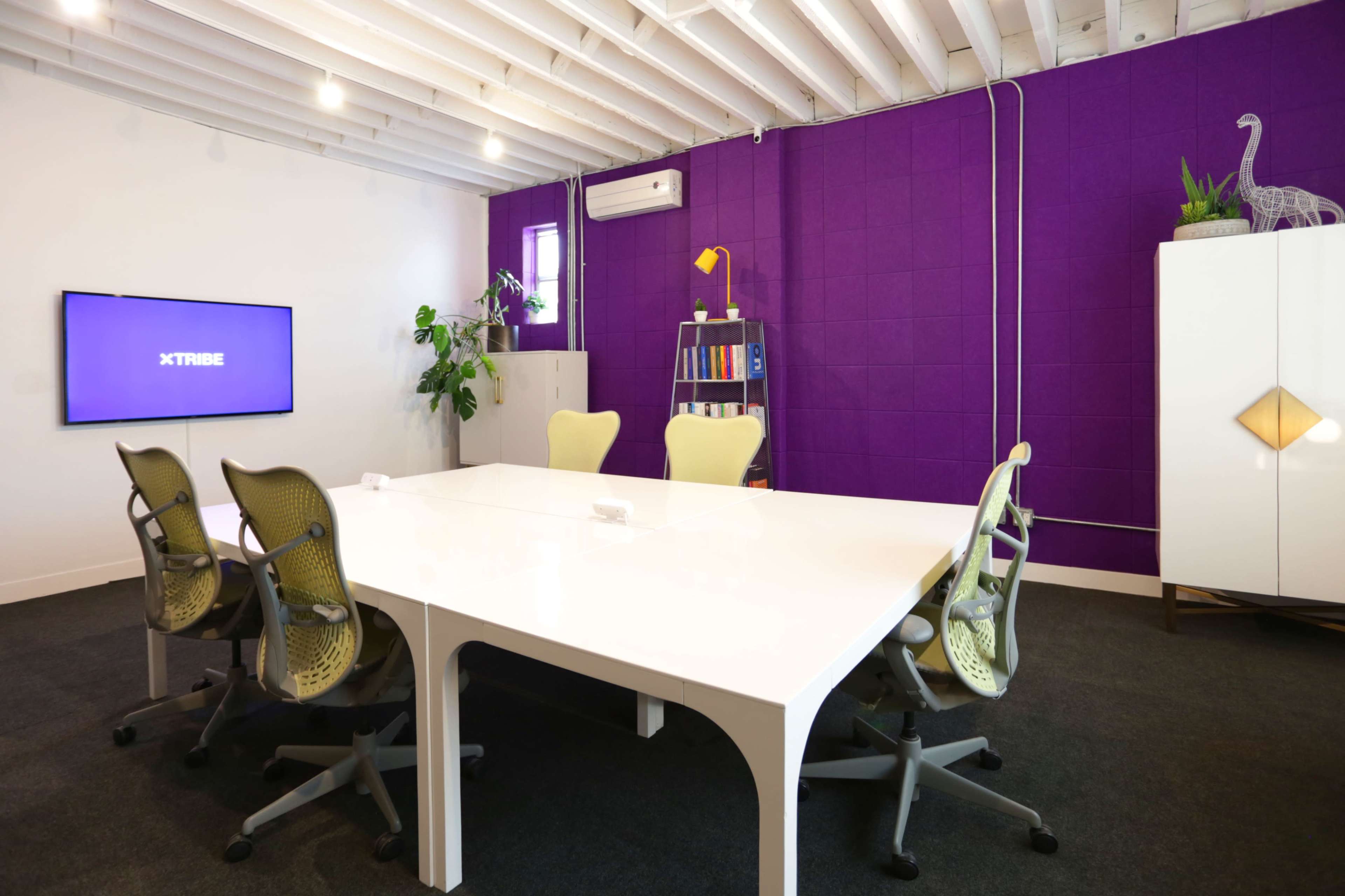 A modern conference room features a large white table surrounded by four ergonomic chairs, with a purple accent wall and a TV screen displaying "XTRIBE."