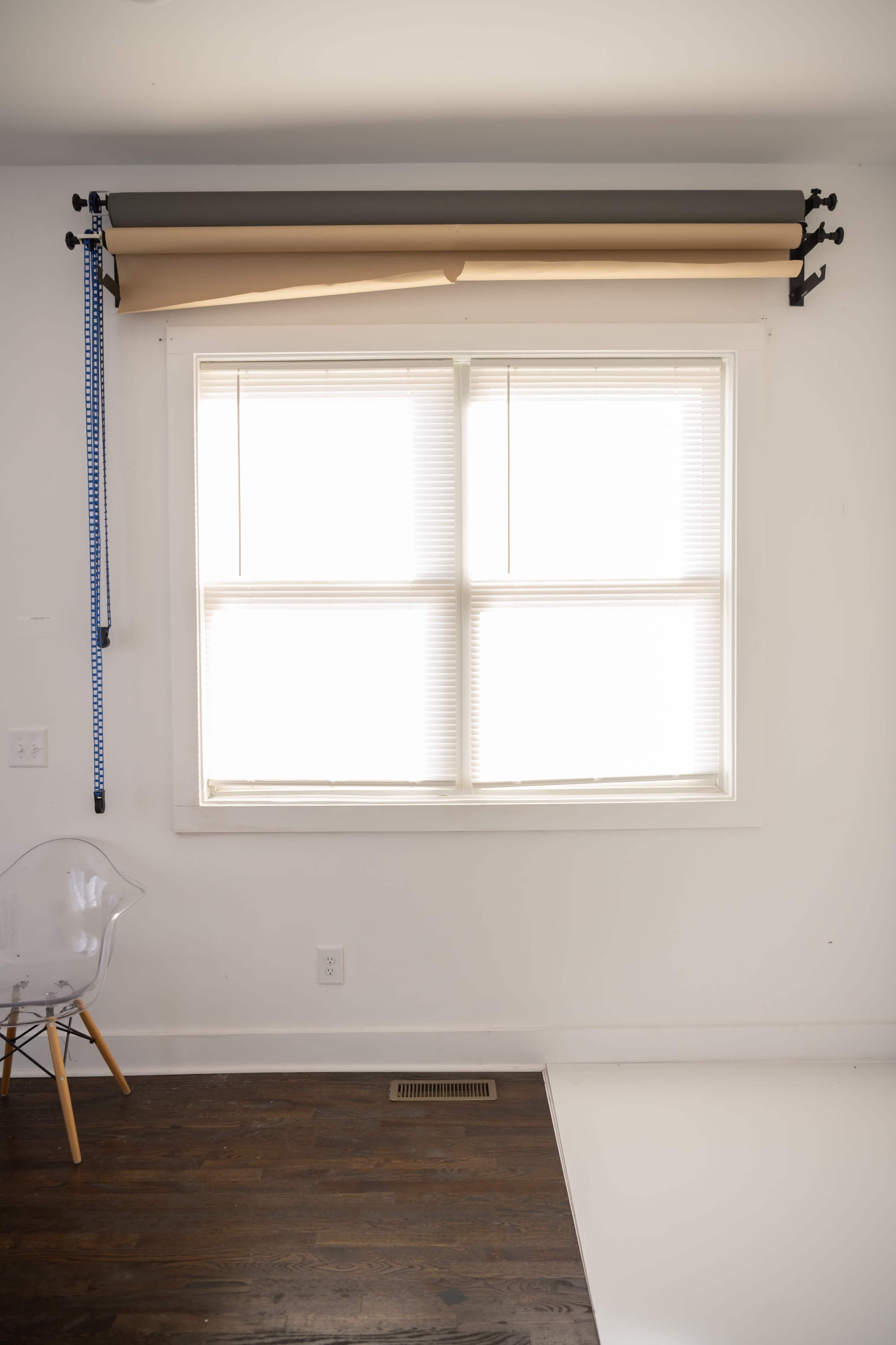 A window with white blinds is located on a white wall, featuring a roll of brown paper and a measuring tape mounted above it.