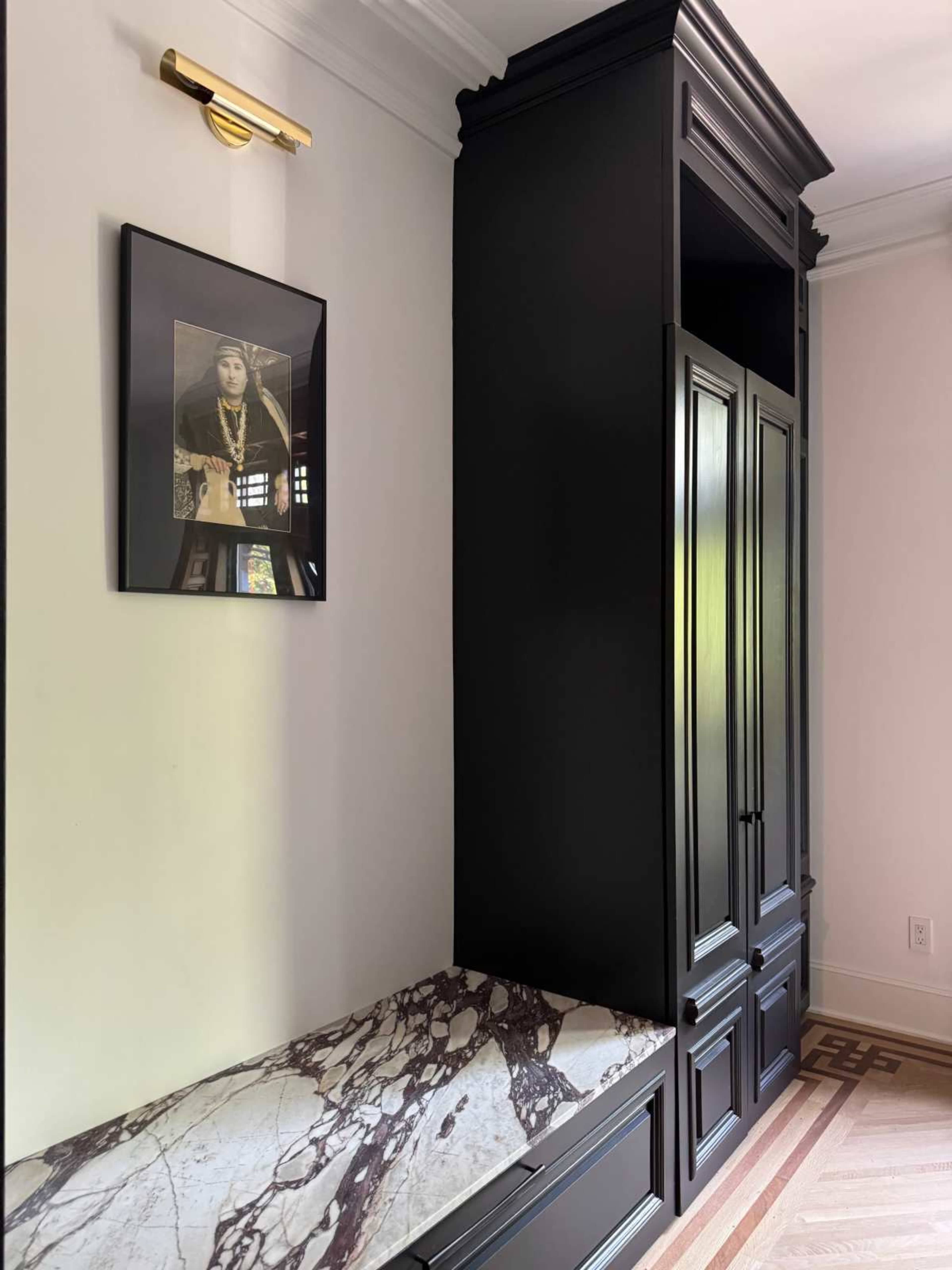 The image shows a corner of a room featuring a sleek black cabinet, a marble-topped bench, and a framed photograph on the wall above.