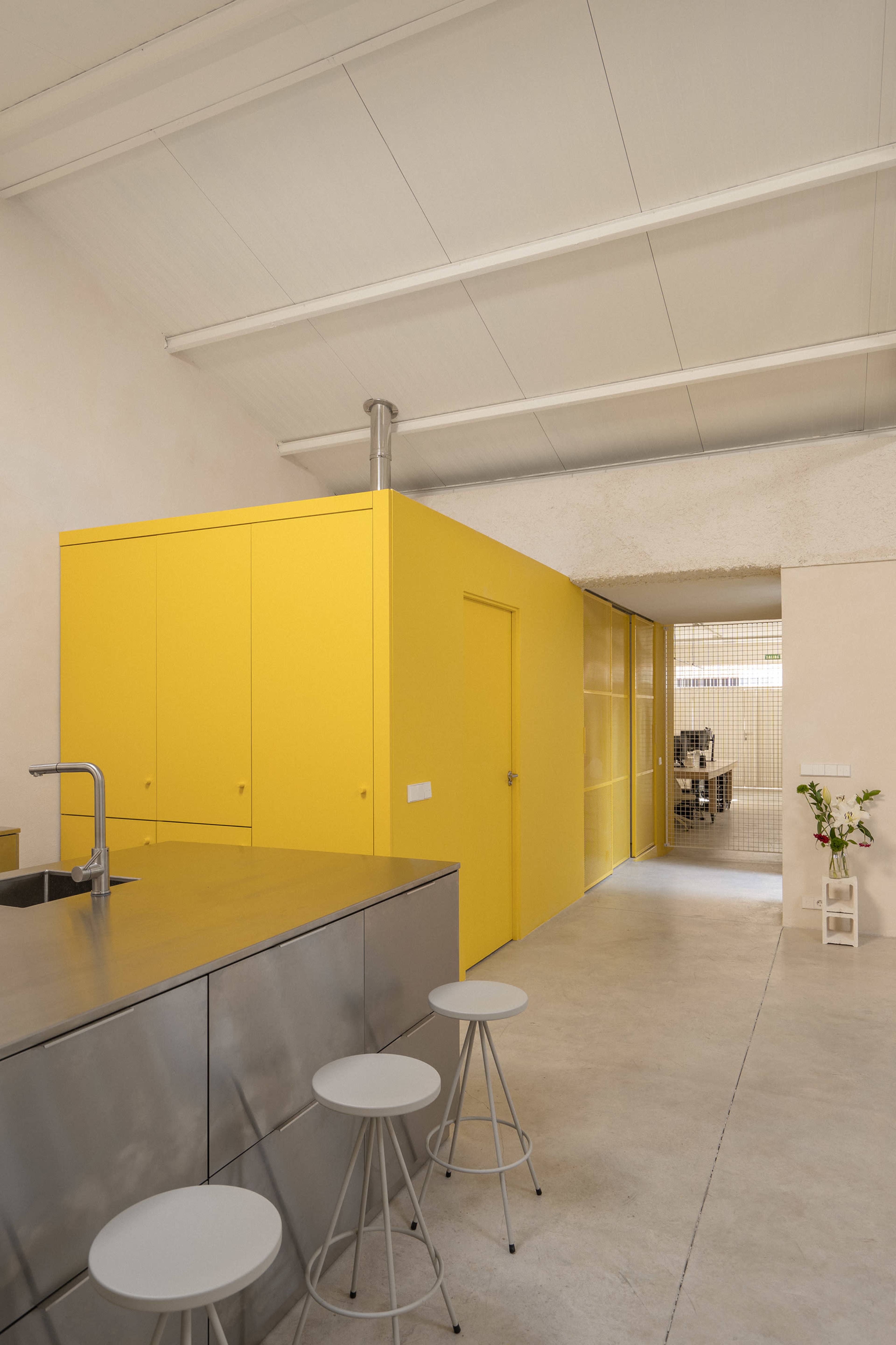 The image shows a modern, minimalist kitchen with a yellow feature wall and a stainless steel island, leading to an interconnected workspace in the background.