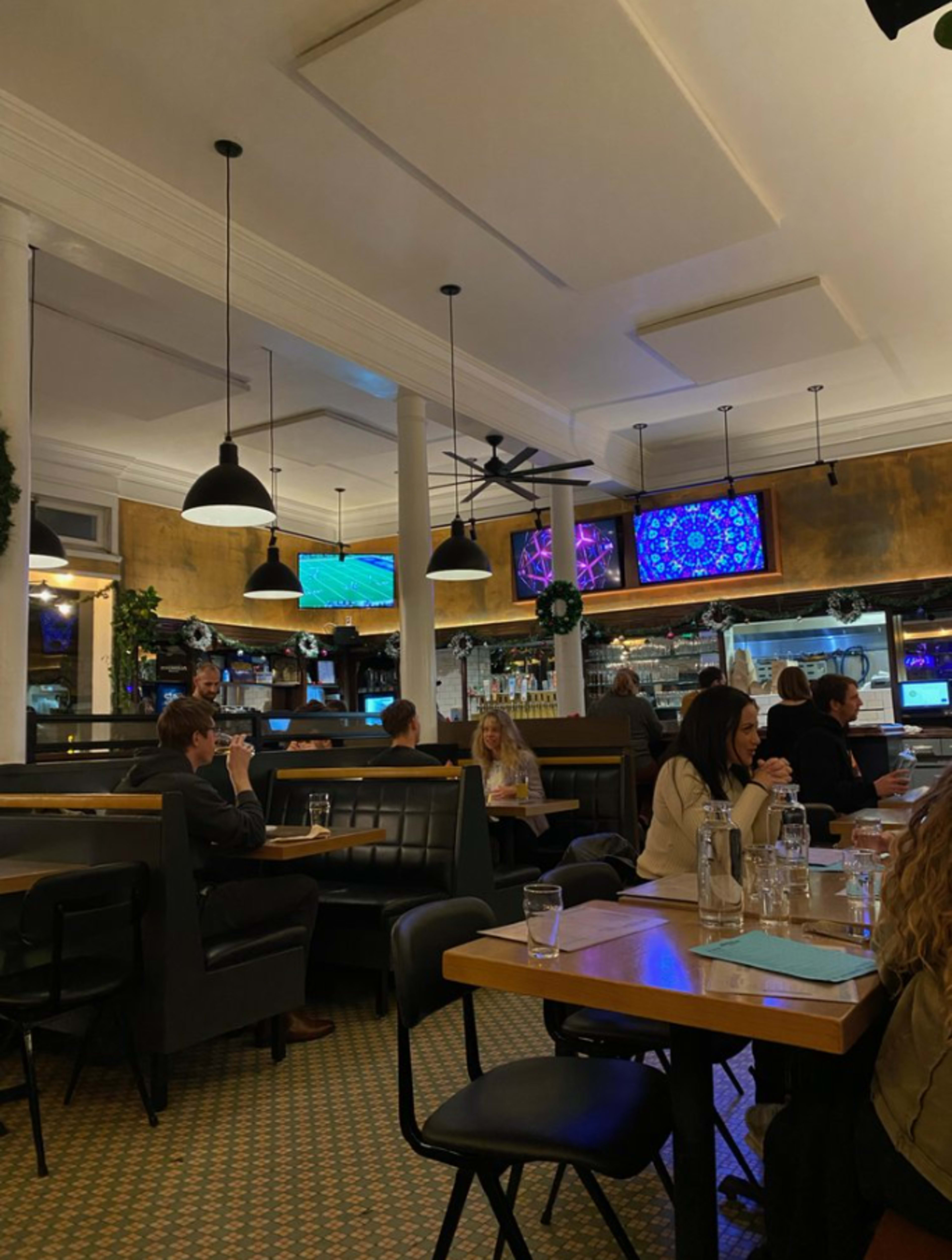 The image shows a spacious restaurant interior with dark seating, several patrons dining, and multiple screens displaying images in a well-lit environment.
