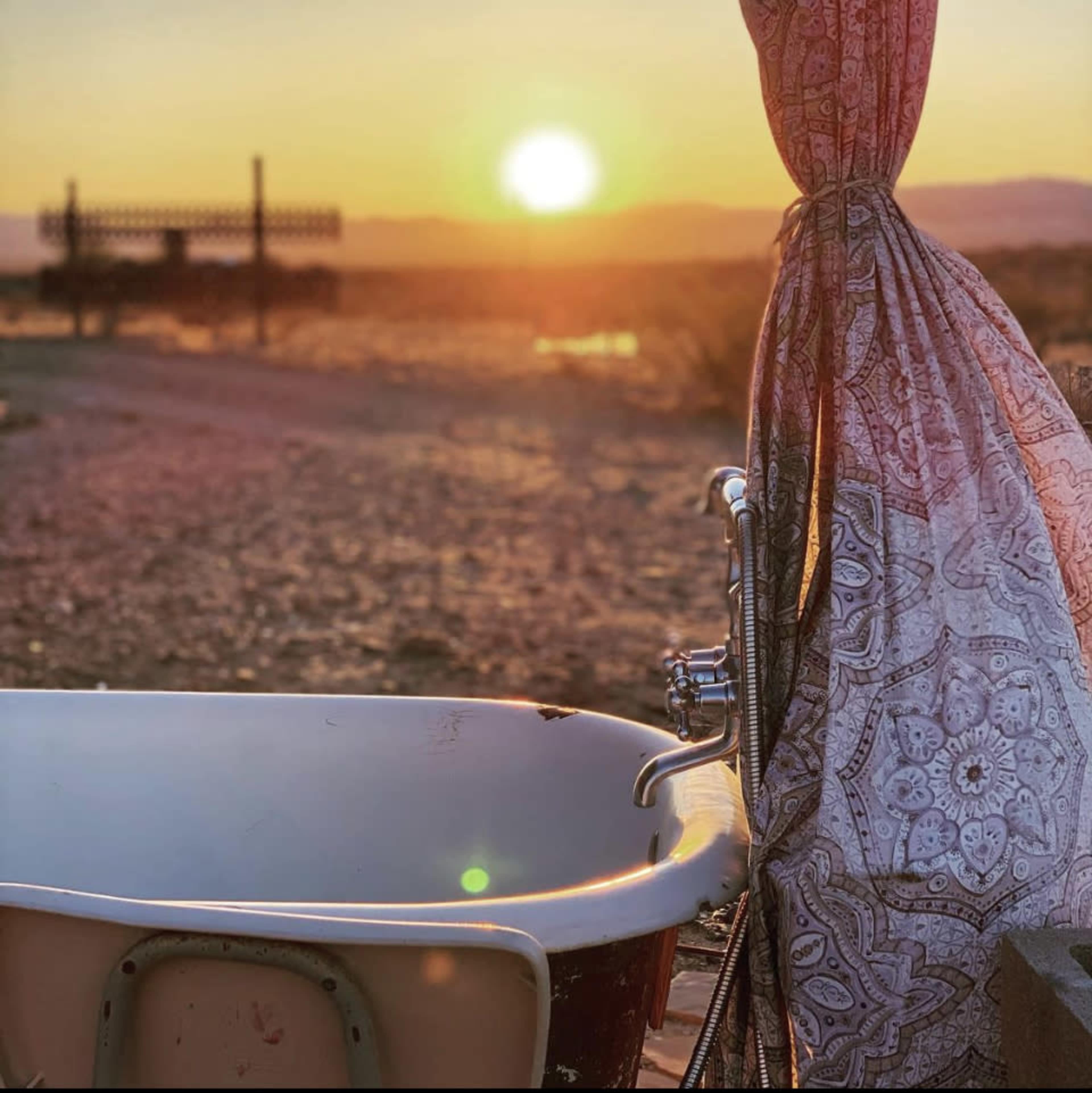 The scene shows a vintage bathtub positioned outdoors near a sunset, with a patterned curtain draped beside it.