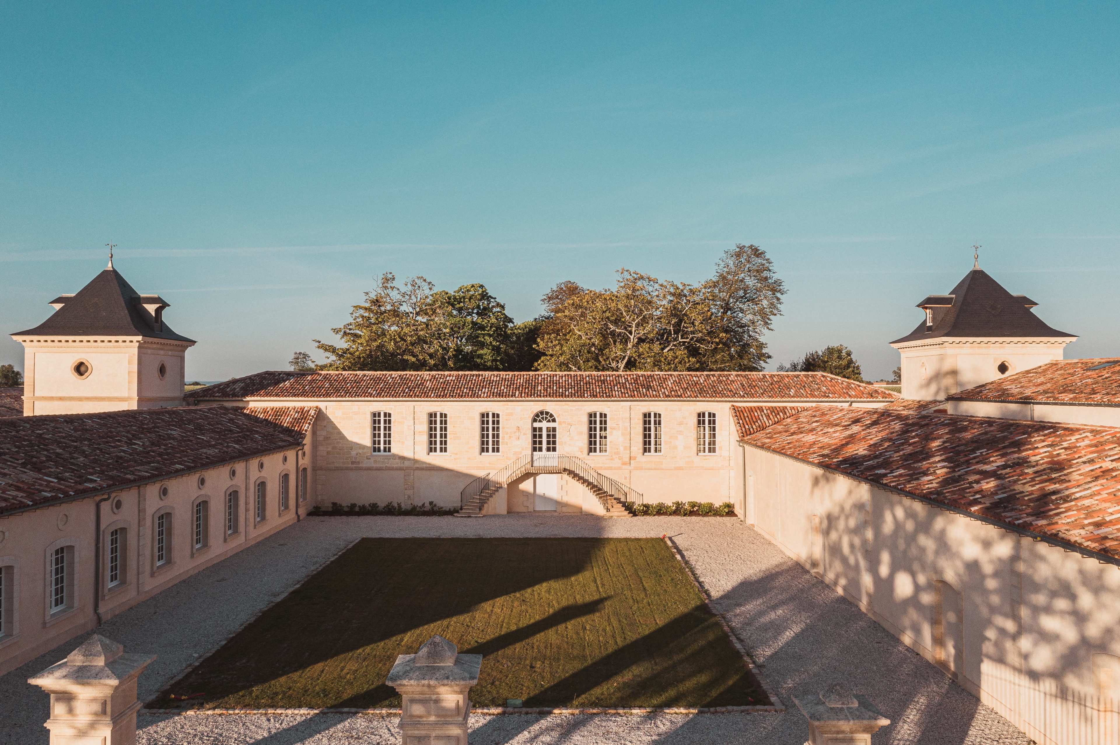 The image shows a symmetrical estate with two tower-like structures flanking a central building, surrounded by a landscaped courtyard and a clear blue sky.