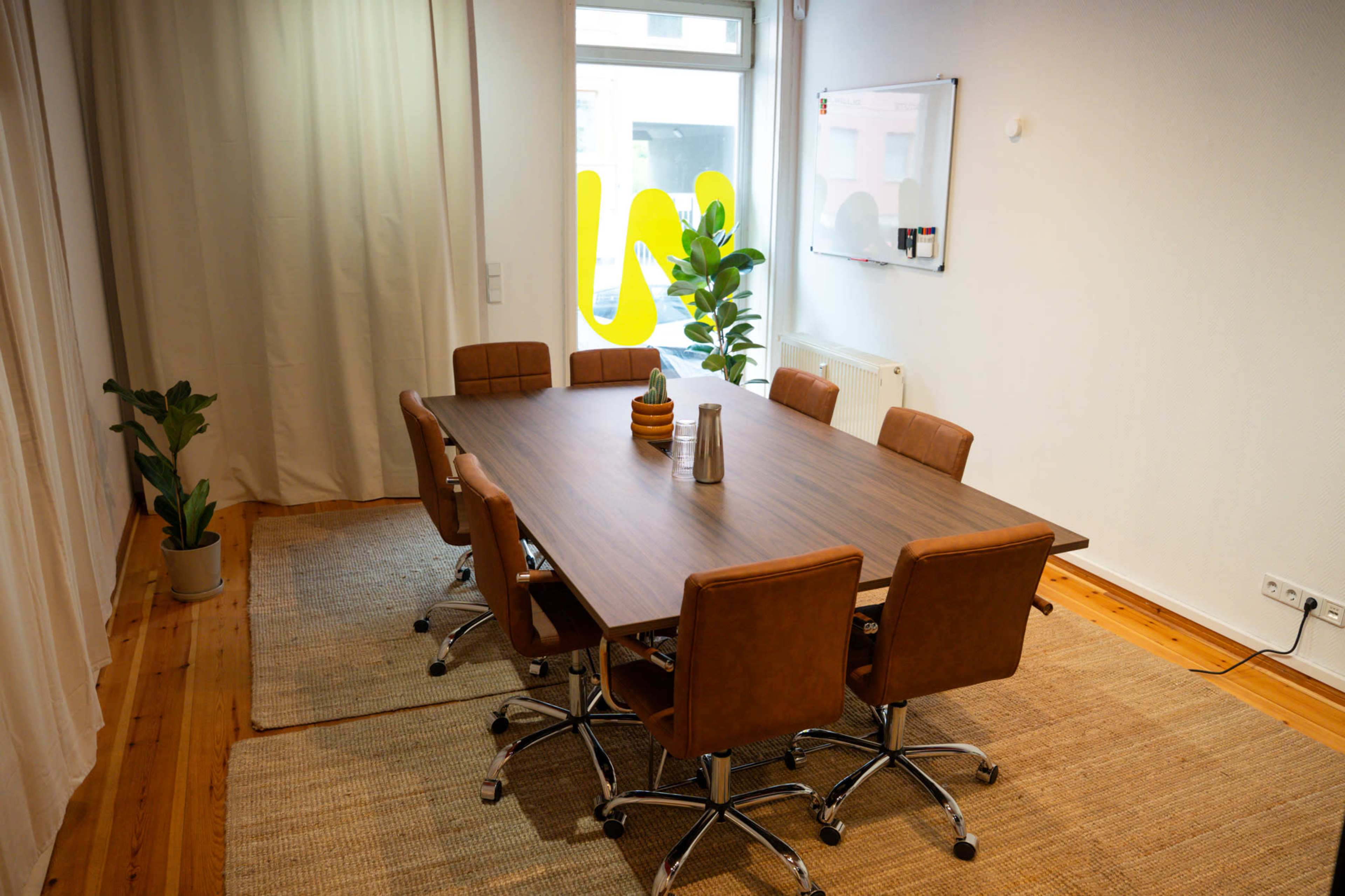 The image shows a modern conference room with a large wooden table surrounded by brown leather chairs, a potted plant in the corner, and natural light streaming in through a window.