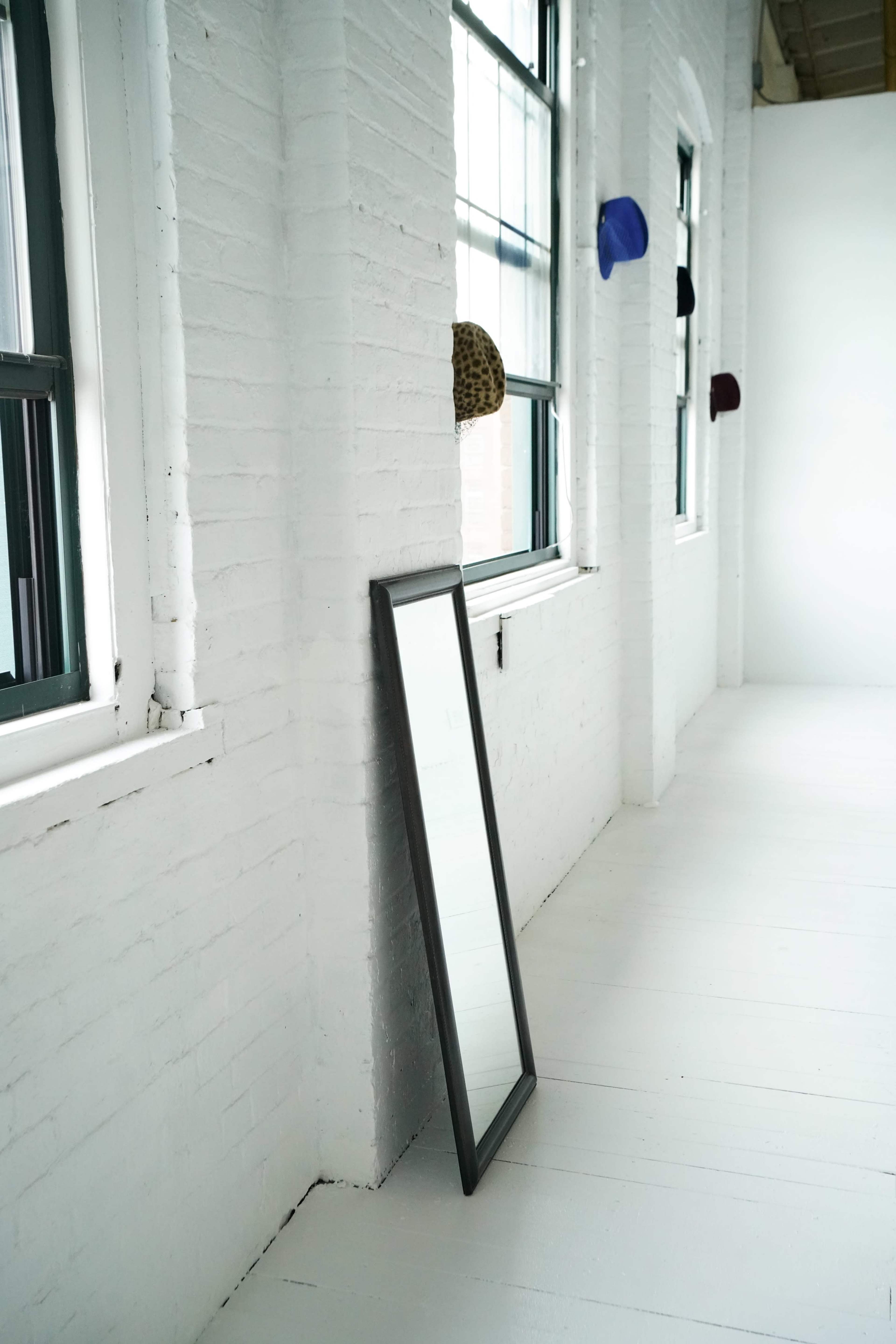 The image shows a bright, minimalistic room with white brick walls, a large mirror propped against the wall, and several hats hanging on the windows.