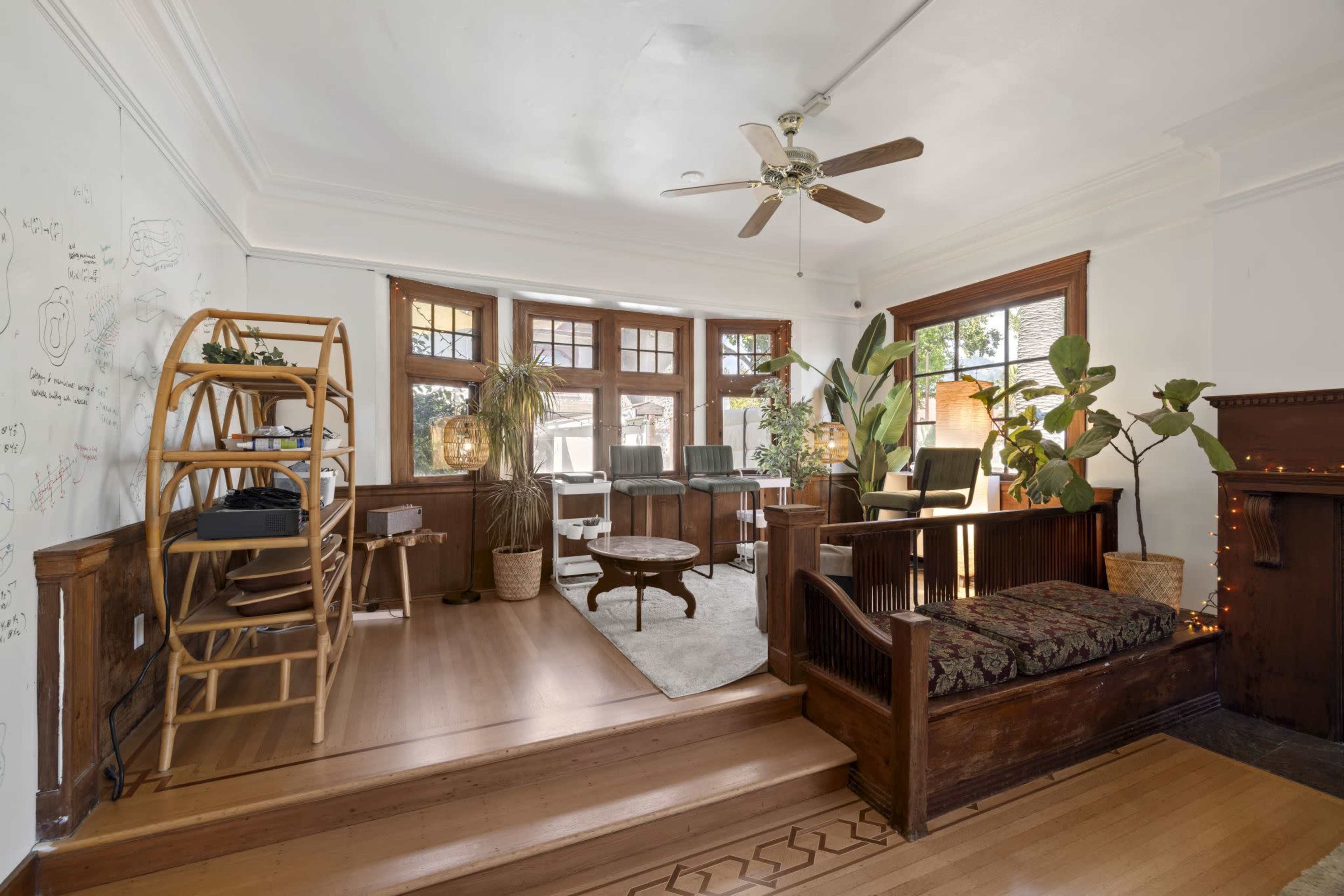 The image depicts a bright and airy living room featuring wooden accents, large windows, and greenery, with seating areas and decorative elements.