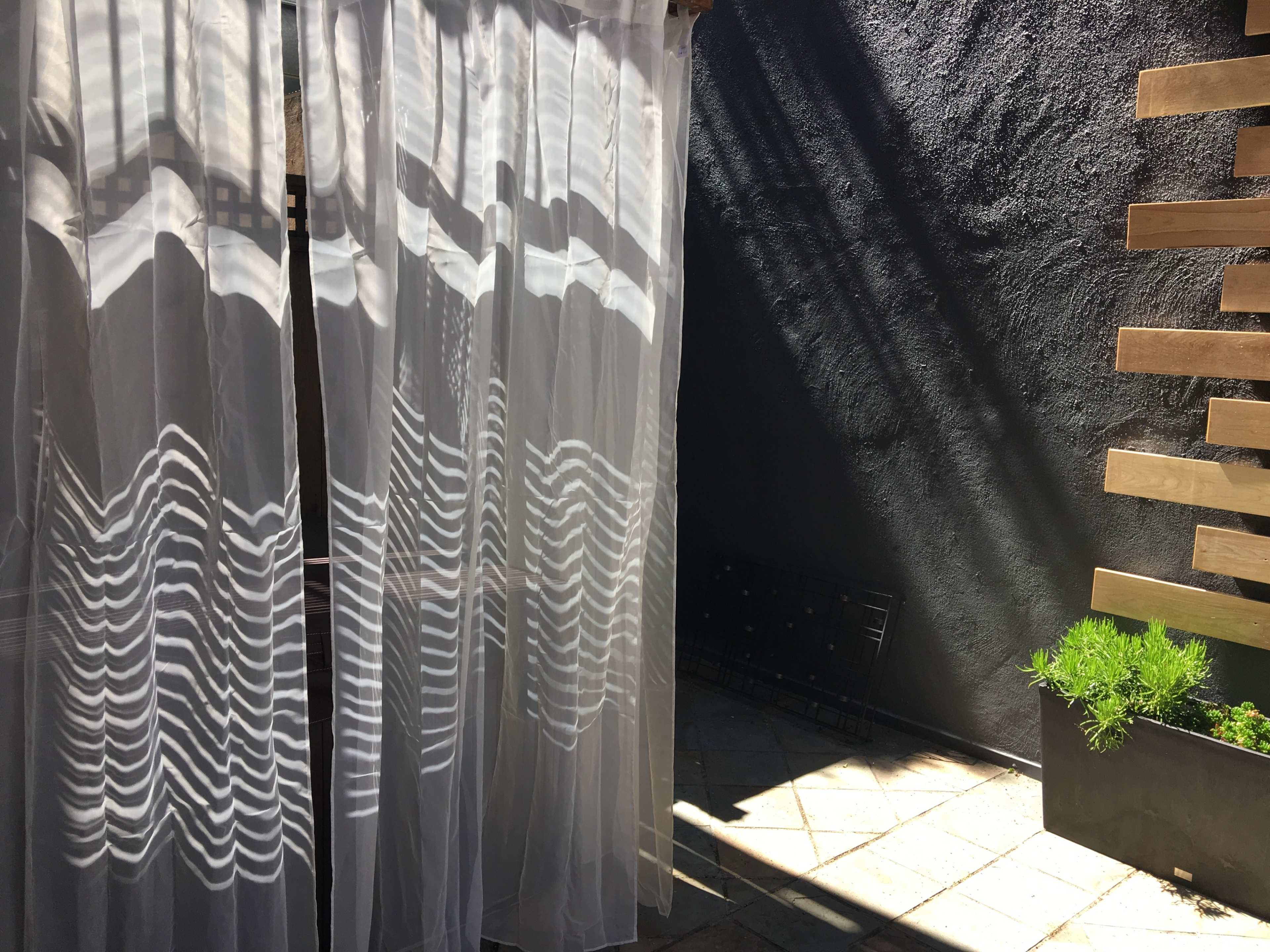 A sheer curtain casts striped shadows on a gray wall beside a planter with greenery.