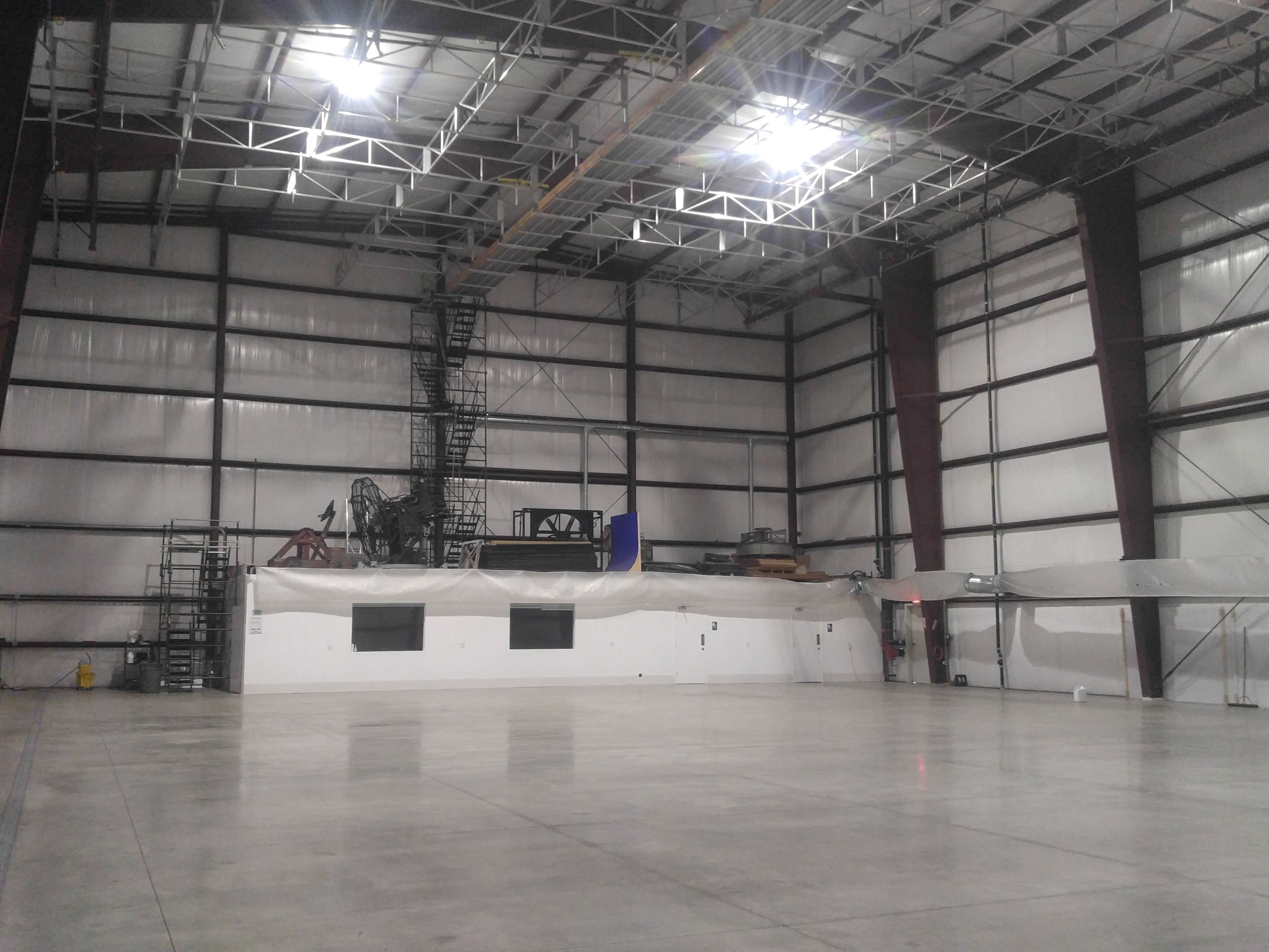 The image shows an expansive industrial warehouse with bare concrete floors and high metal ceilings, featuring a staircase and machinery against one wall.
