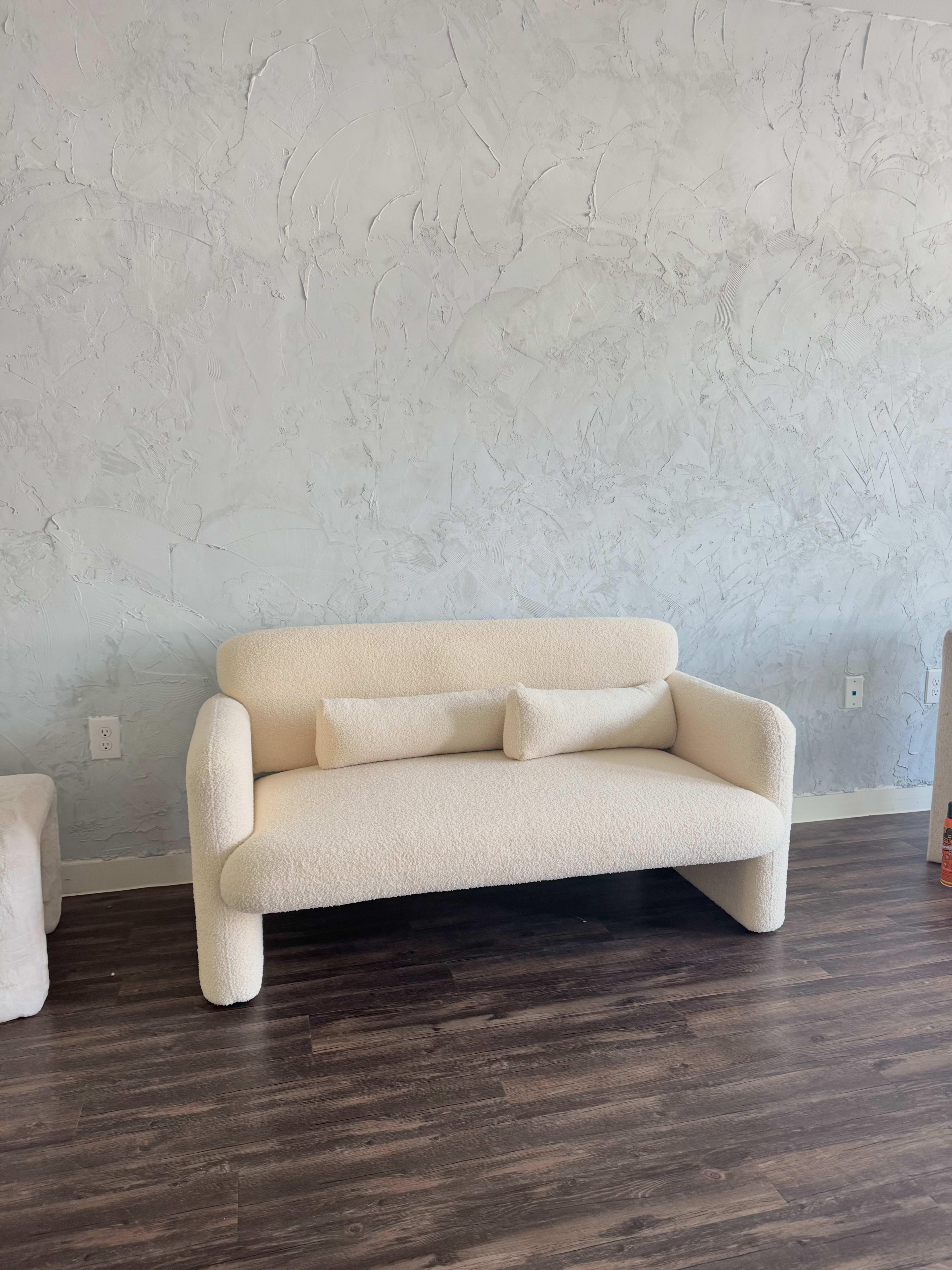 The image features a cream-colored, modern sofa with a minimalist design, set against a textured gray wall and wood flooring.
