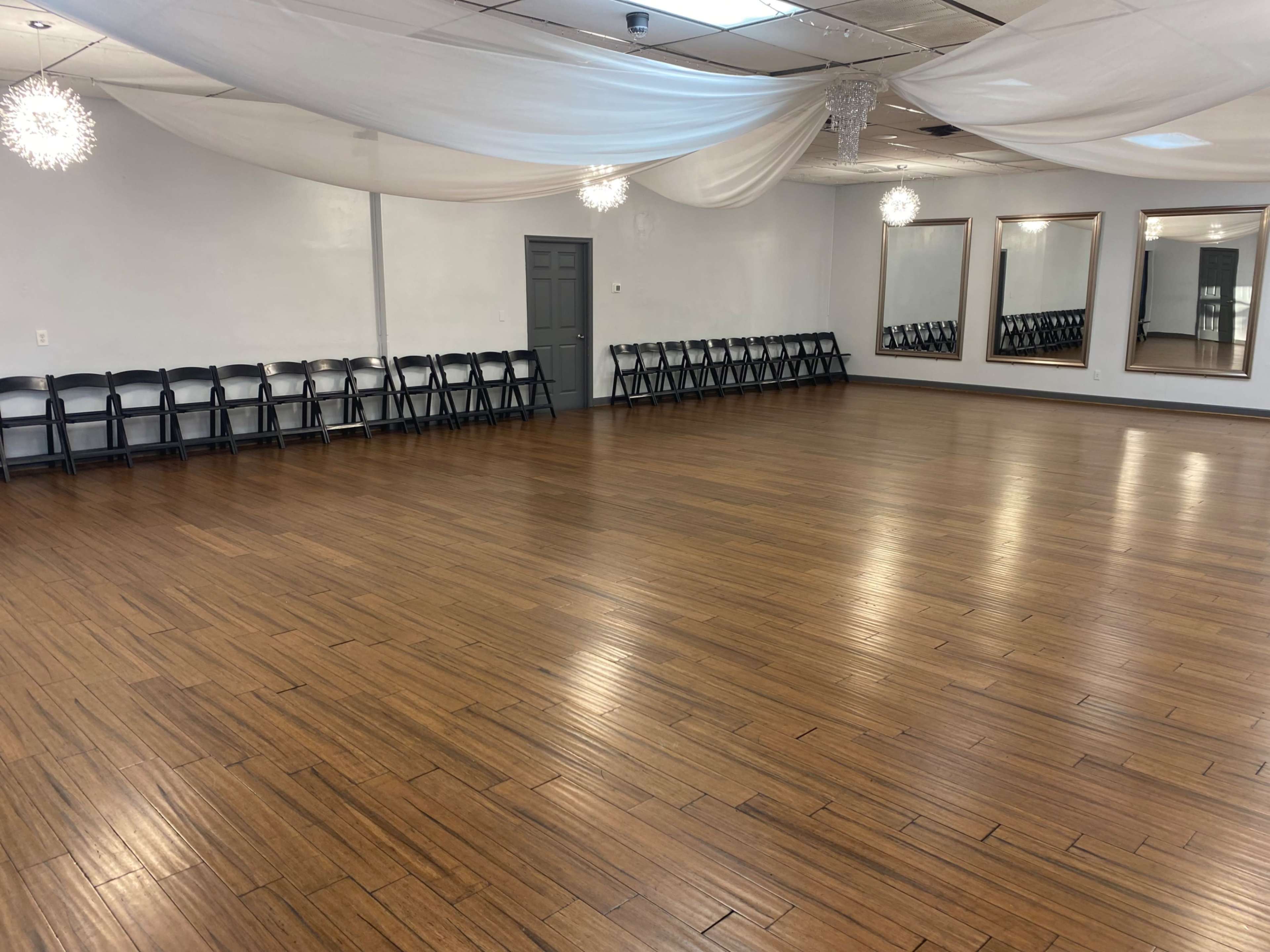 An empty wooden floor ballroom with black folding chairs arranged along one side and decorative mirrors on the opposite wall.