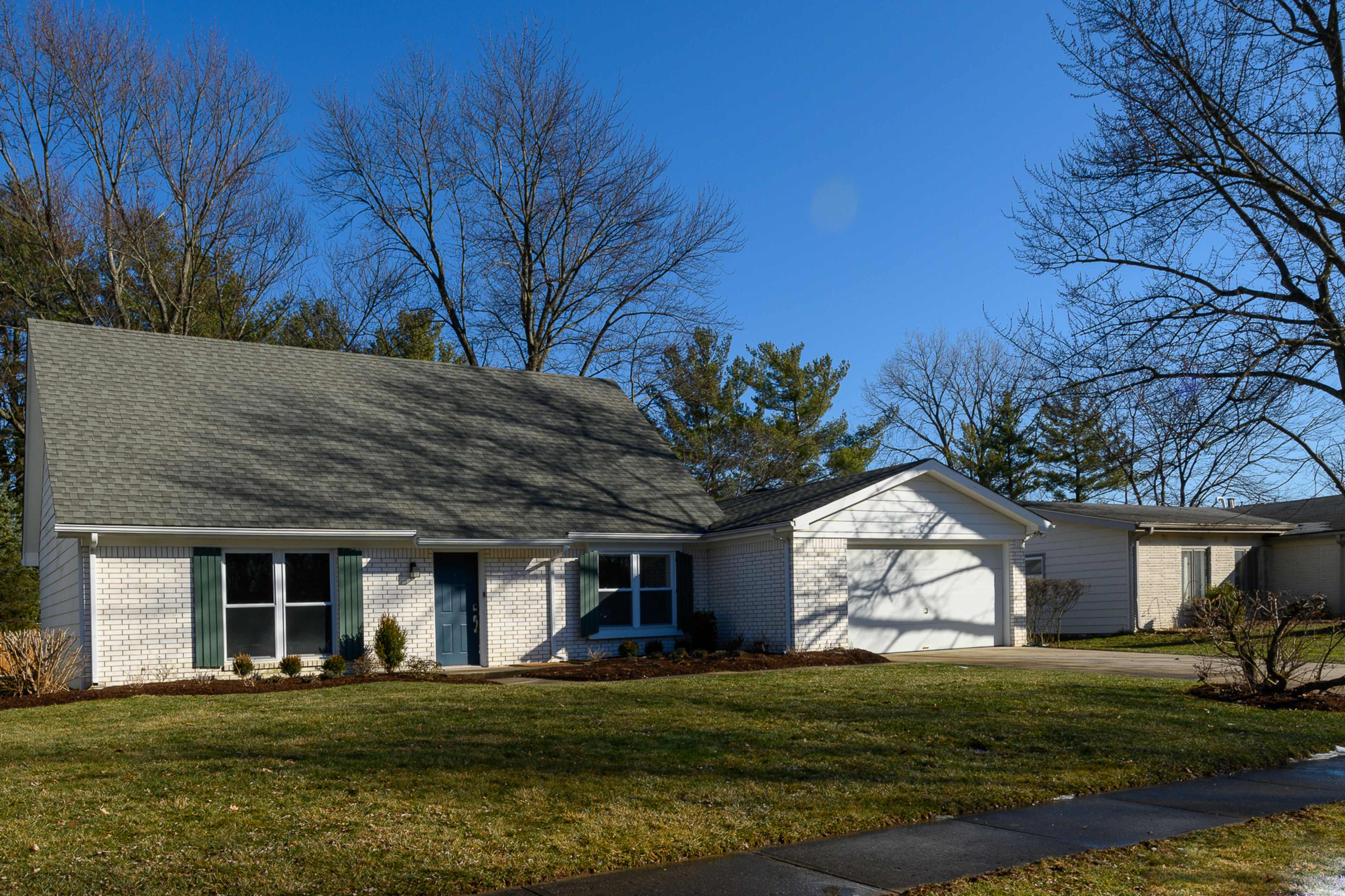 A single-story house with a gabled roof, green shutters, and a two-car garage is situated on a lawn surrounded by trees.