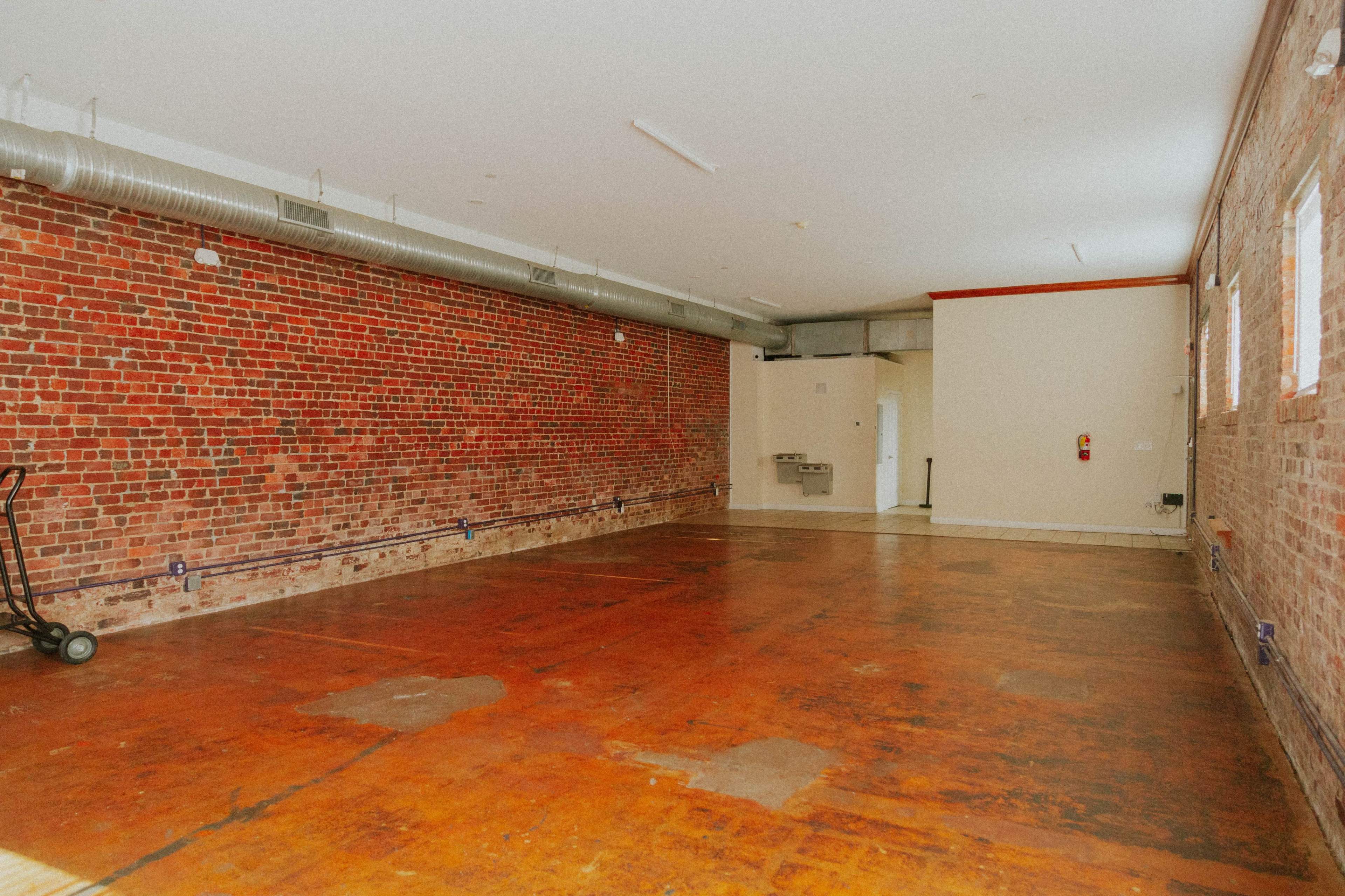 The image shows a large, empty room with exposed brick walls and a wooden floor, featuring a utility sink in one corner.