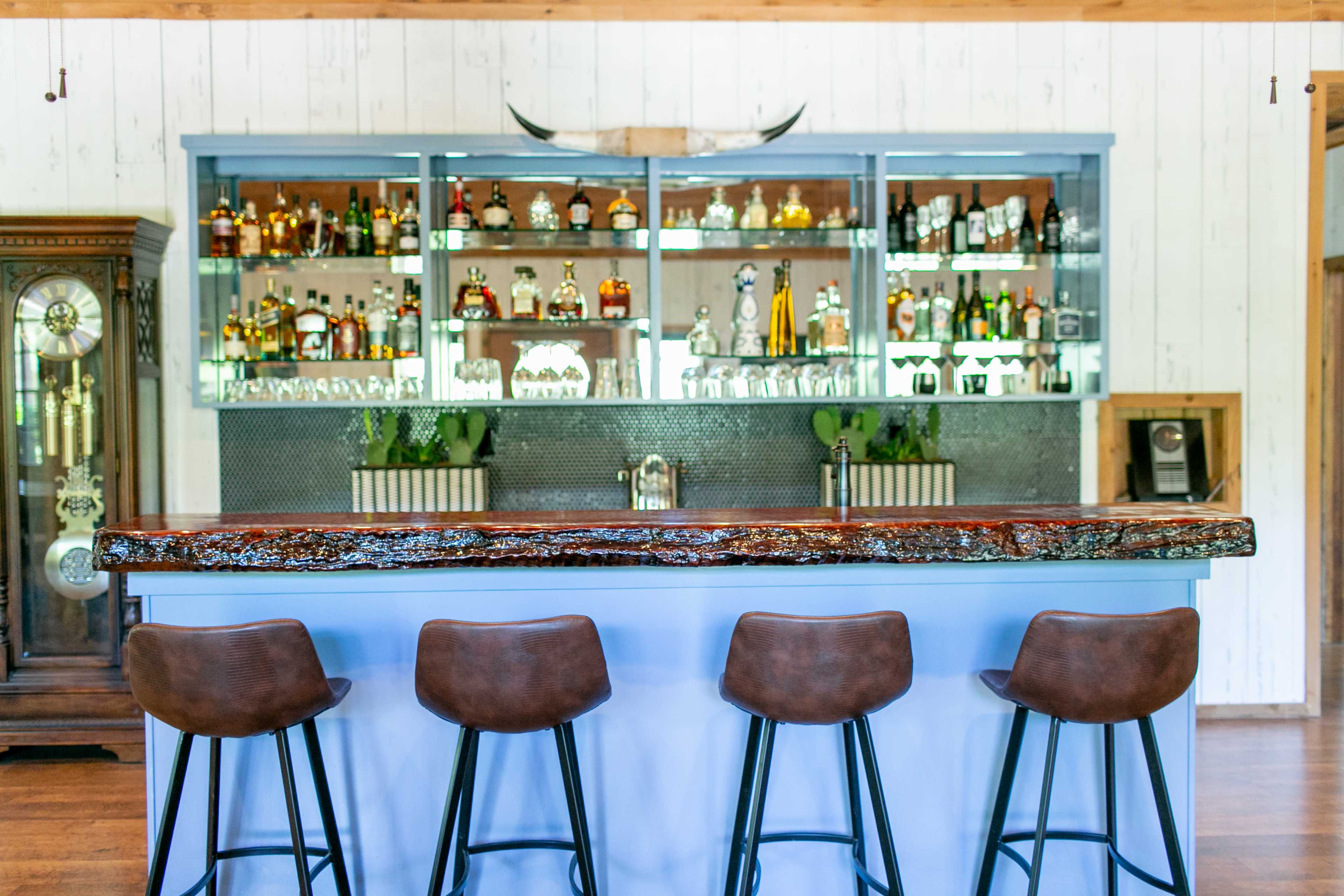 The image shows a modern bar with a wooden counter and four brown stools, set against a backdrop of glass shelves displaying various bottles of liquor and a rustic decor.