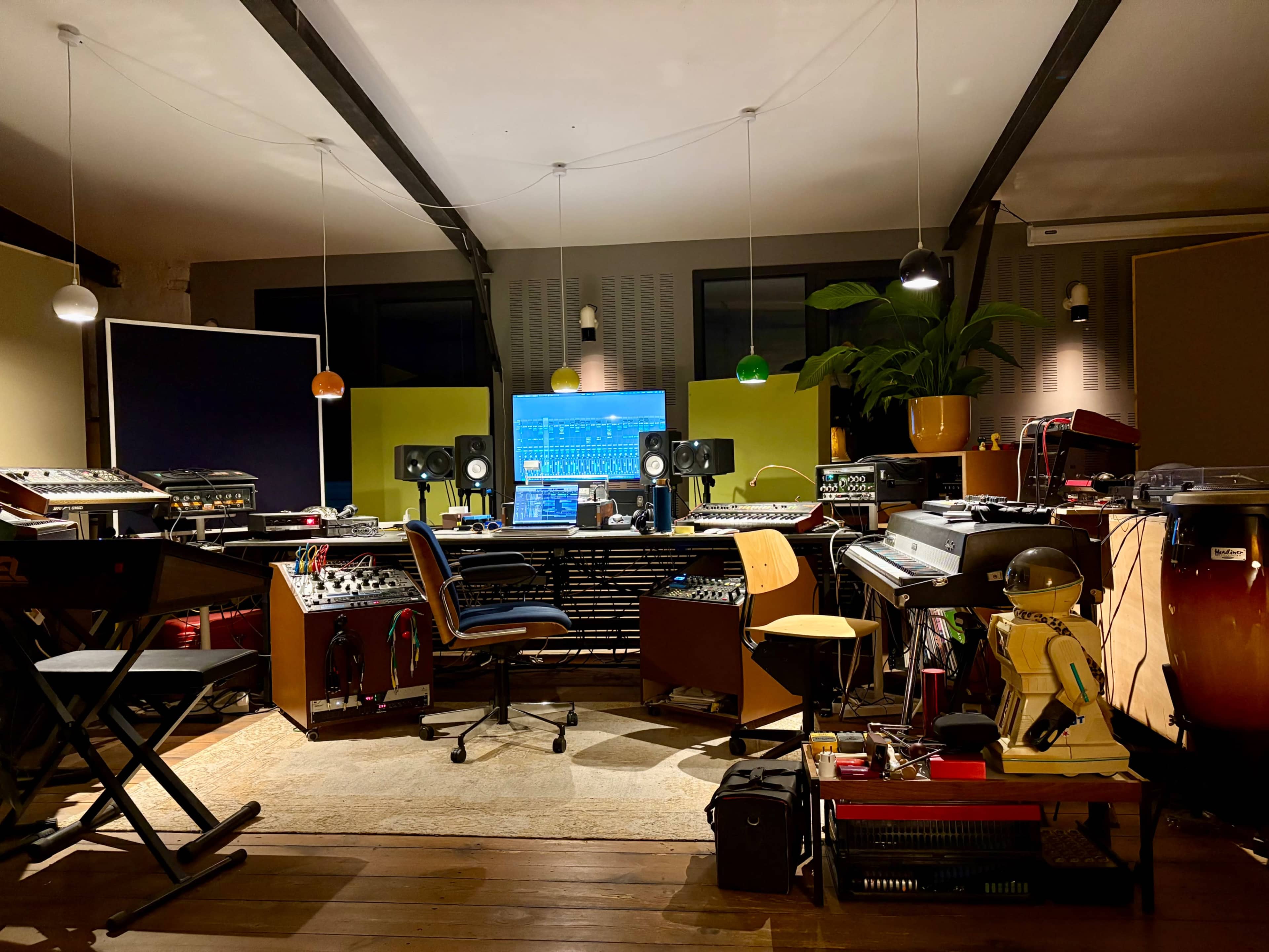 The image shows a music studio filled with various instruments, equipment, and a large computer screen displaying a digital audio workstation.