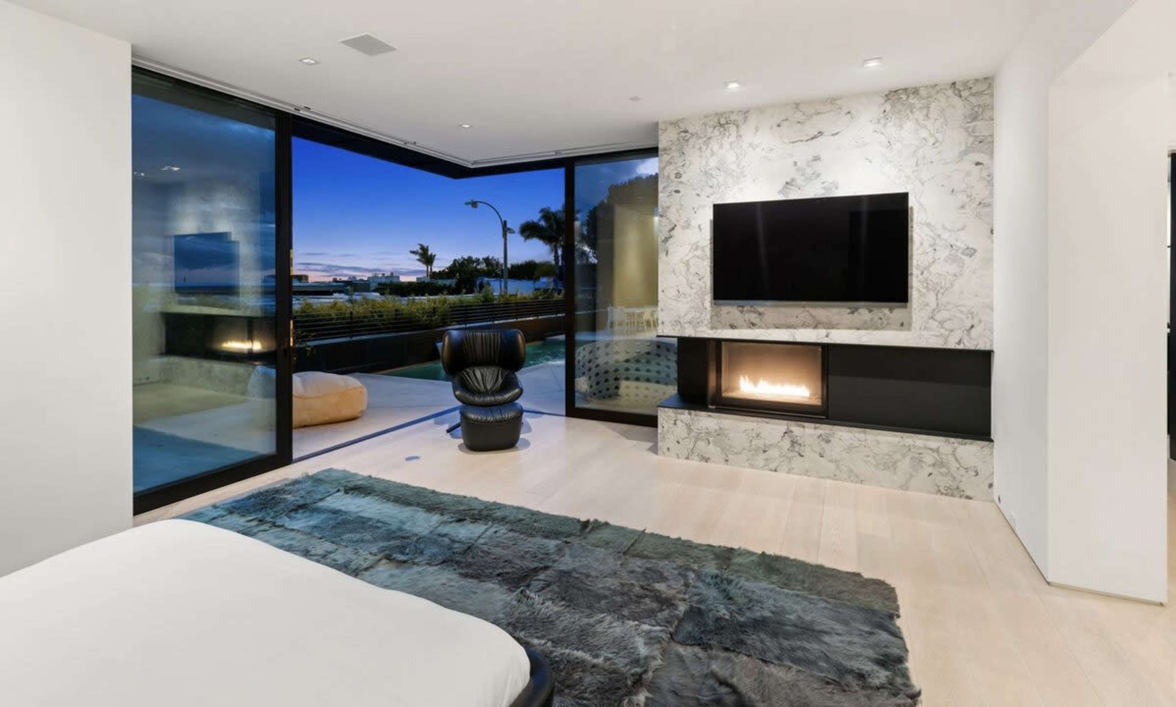 A modern bedroom features a large window with a view, a wall-mounted television, and a fireplace with marble accents.