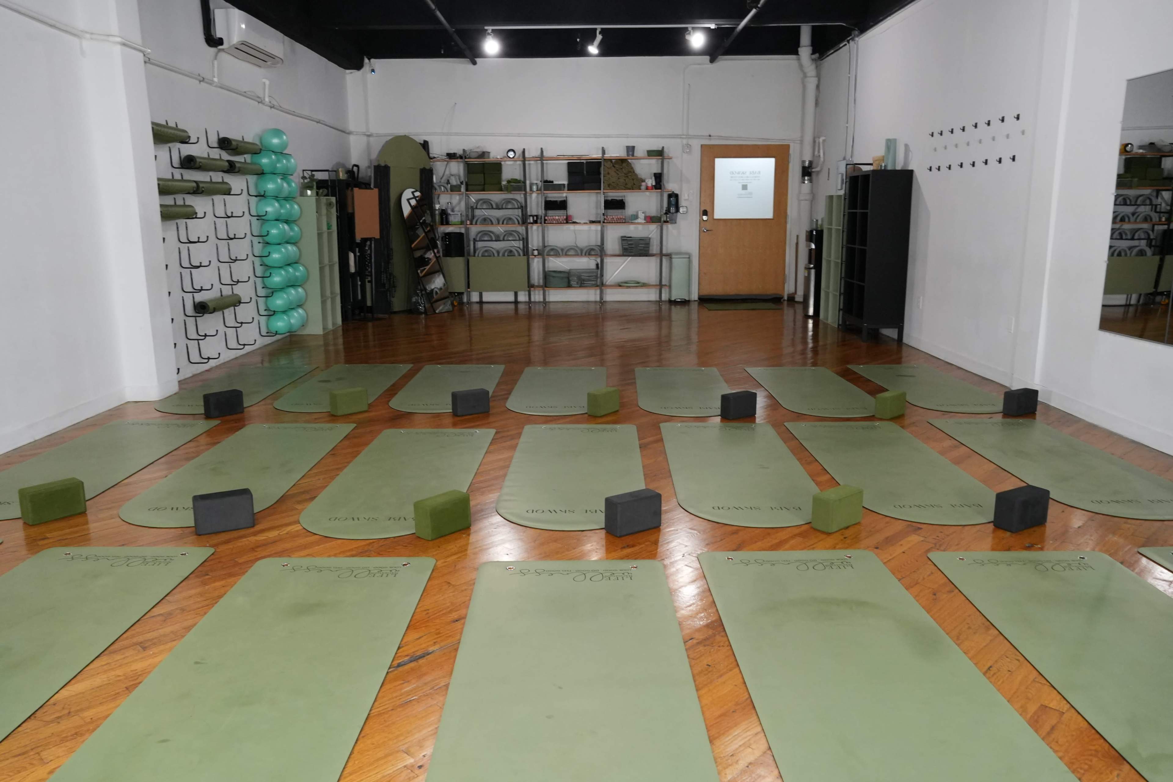 The image shows a yoga studio with green mats arranged in rows on a wooden floor, accompanied by yoga blocks and exercise equipment in the background.