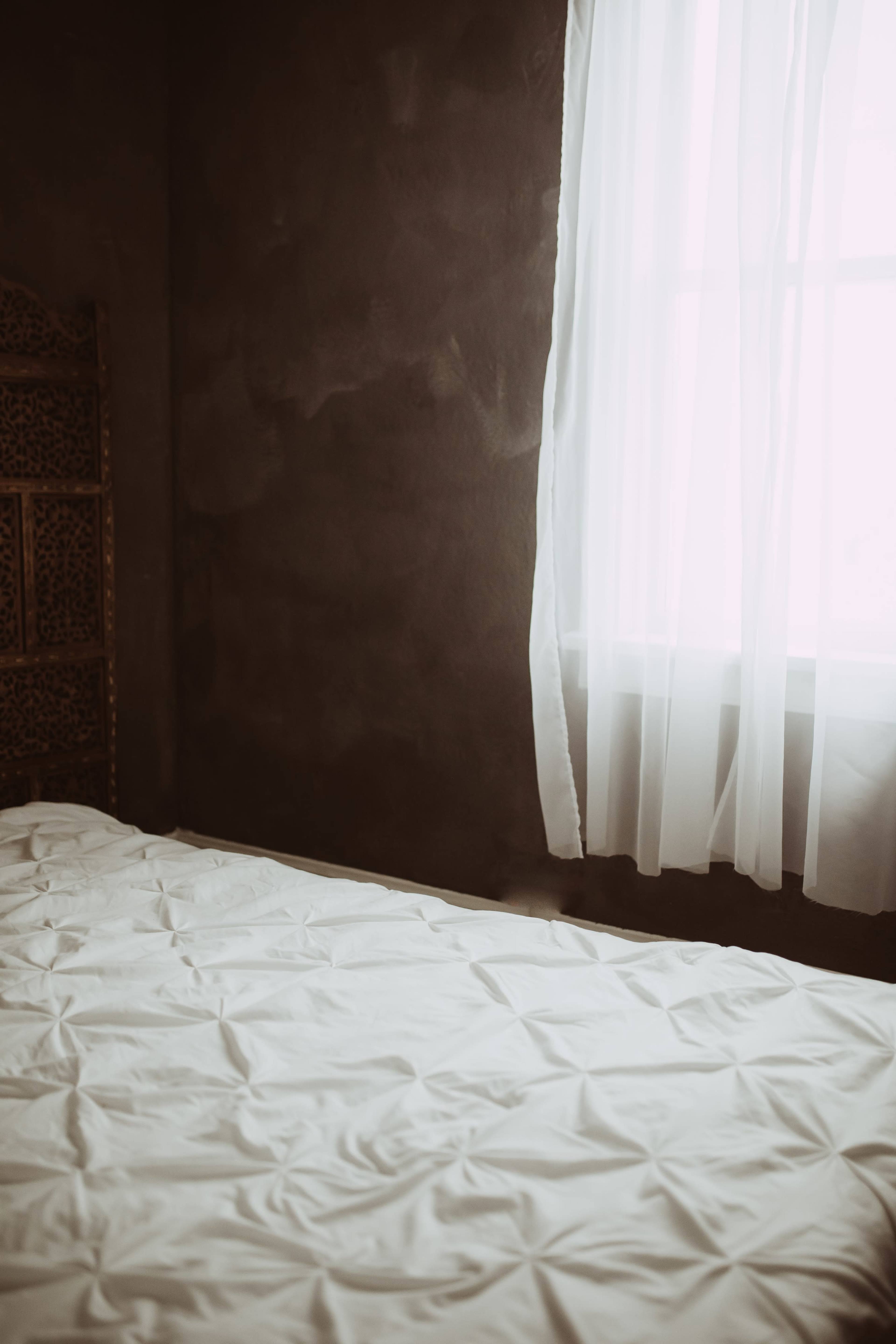 A plain white bed with crumpled sheets is positioned next to a window covered by a sheer curtain in a dim room.