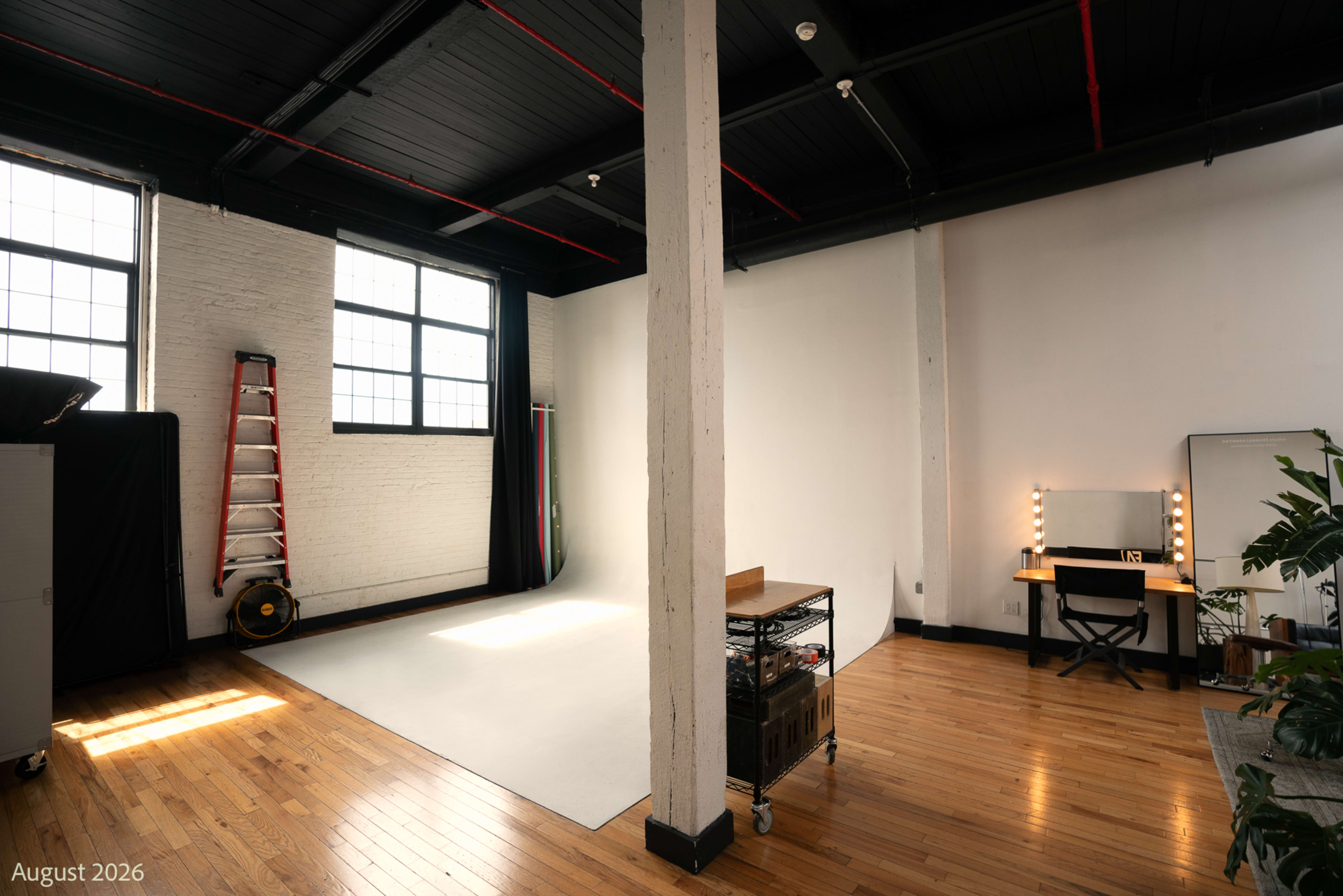 The image shows a bright, spacious studio with a white backdrop, a seating area, and a vanity mirror along with wooden flooring and a tall ladder in the corner.