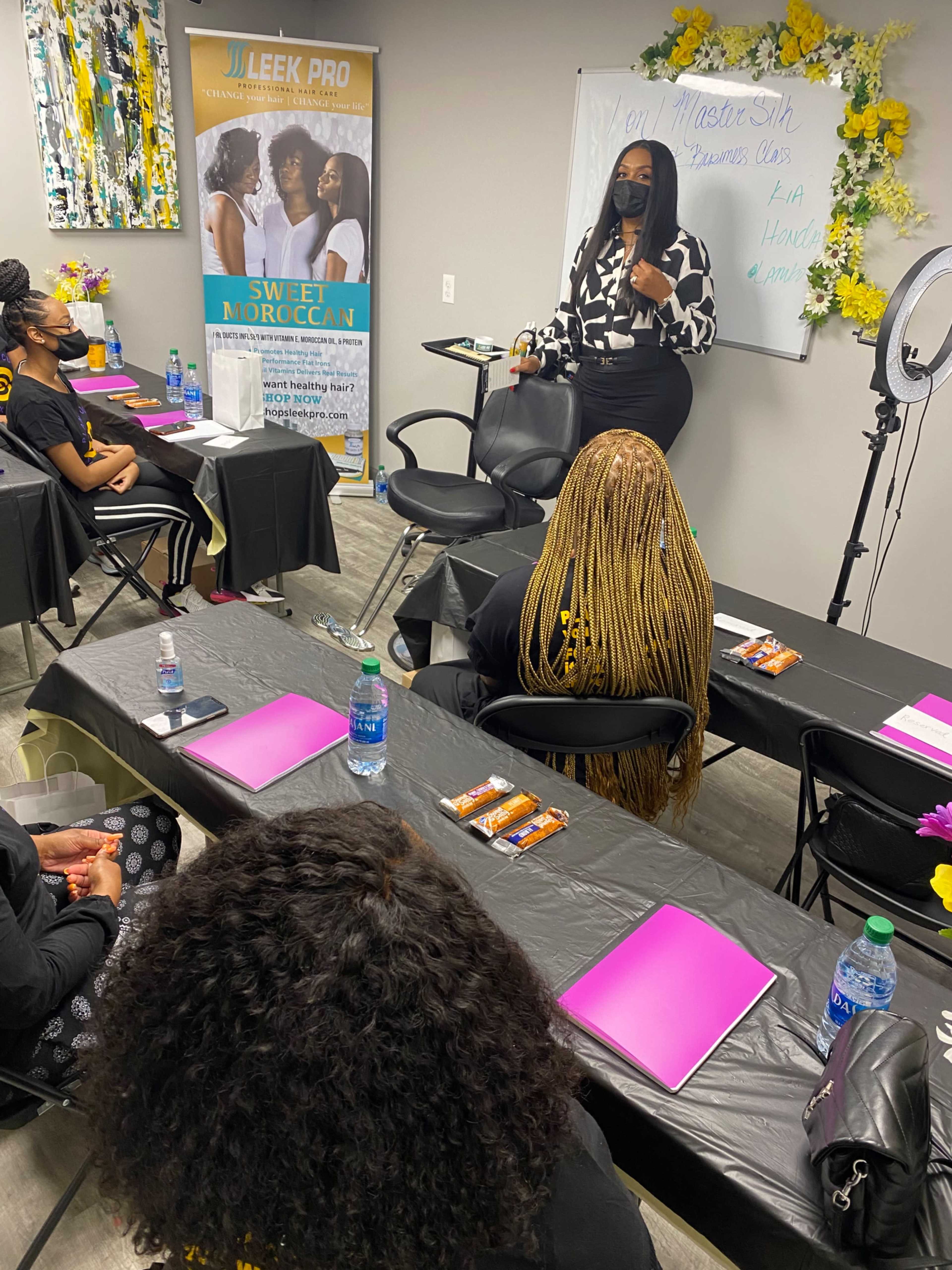 A woman stands in front of a classroom, addressing seated attendees who are facing her, while a whiteboard and promotional banners are visible in the background.