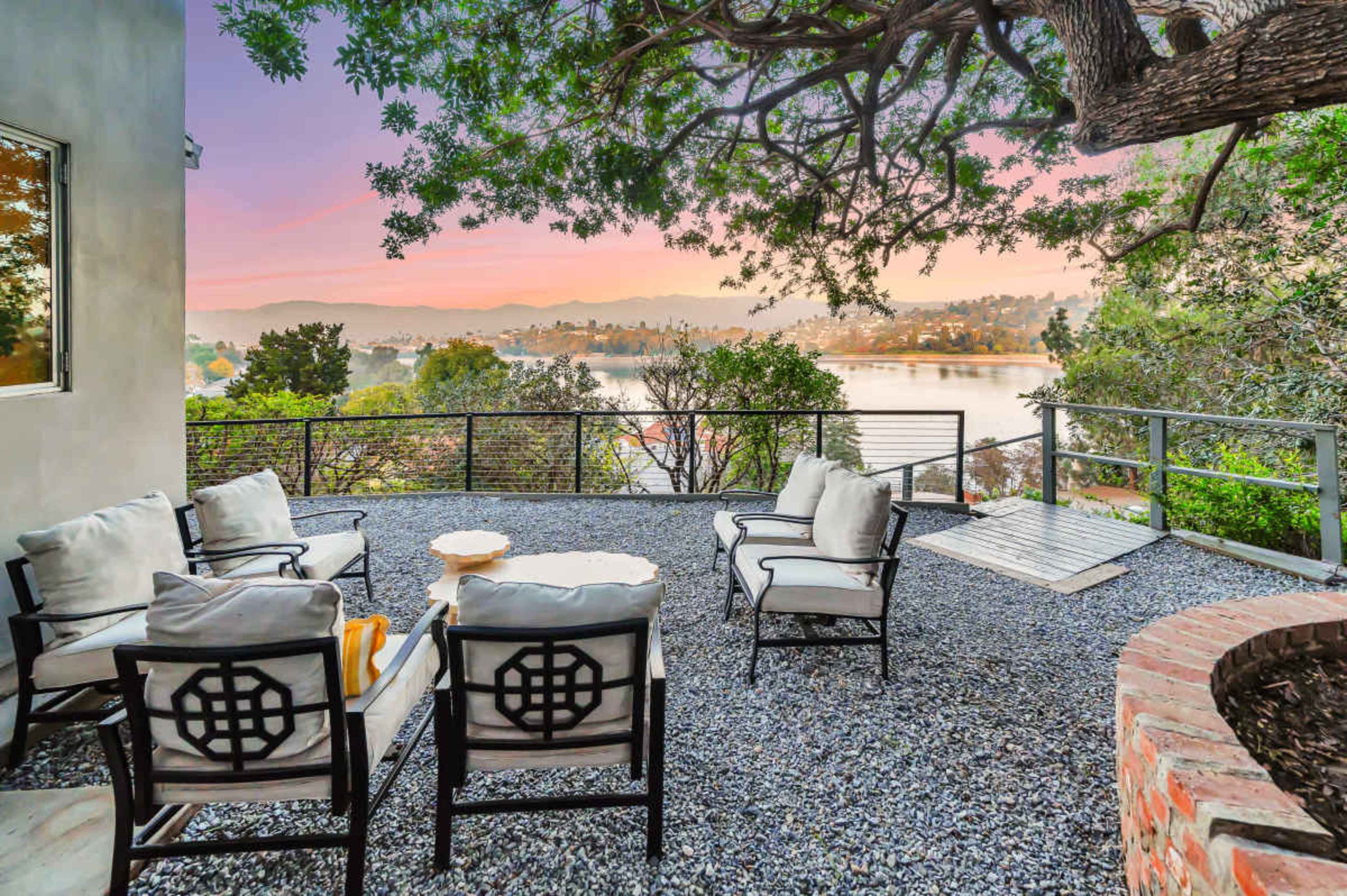 A patio with seating furniture overlooks a body of water and surrounding landscape at sunset.