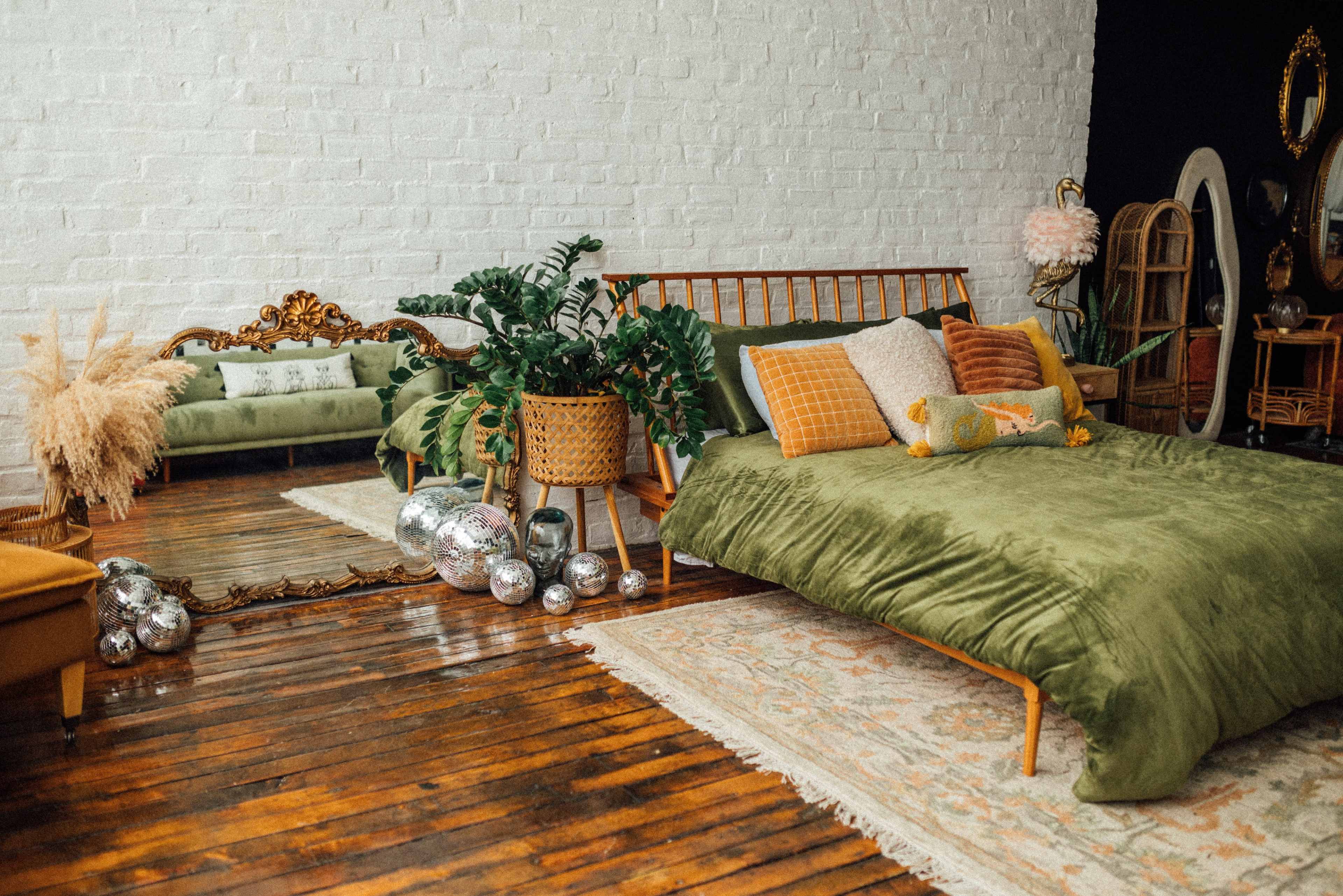 The scene features a cozy bedroom with a green bedding set, a wooden bed frame, a vintage-style mirror, and decorative plants, all arranged on a wooden floor with a patterned rug.