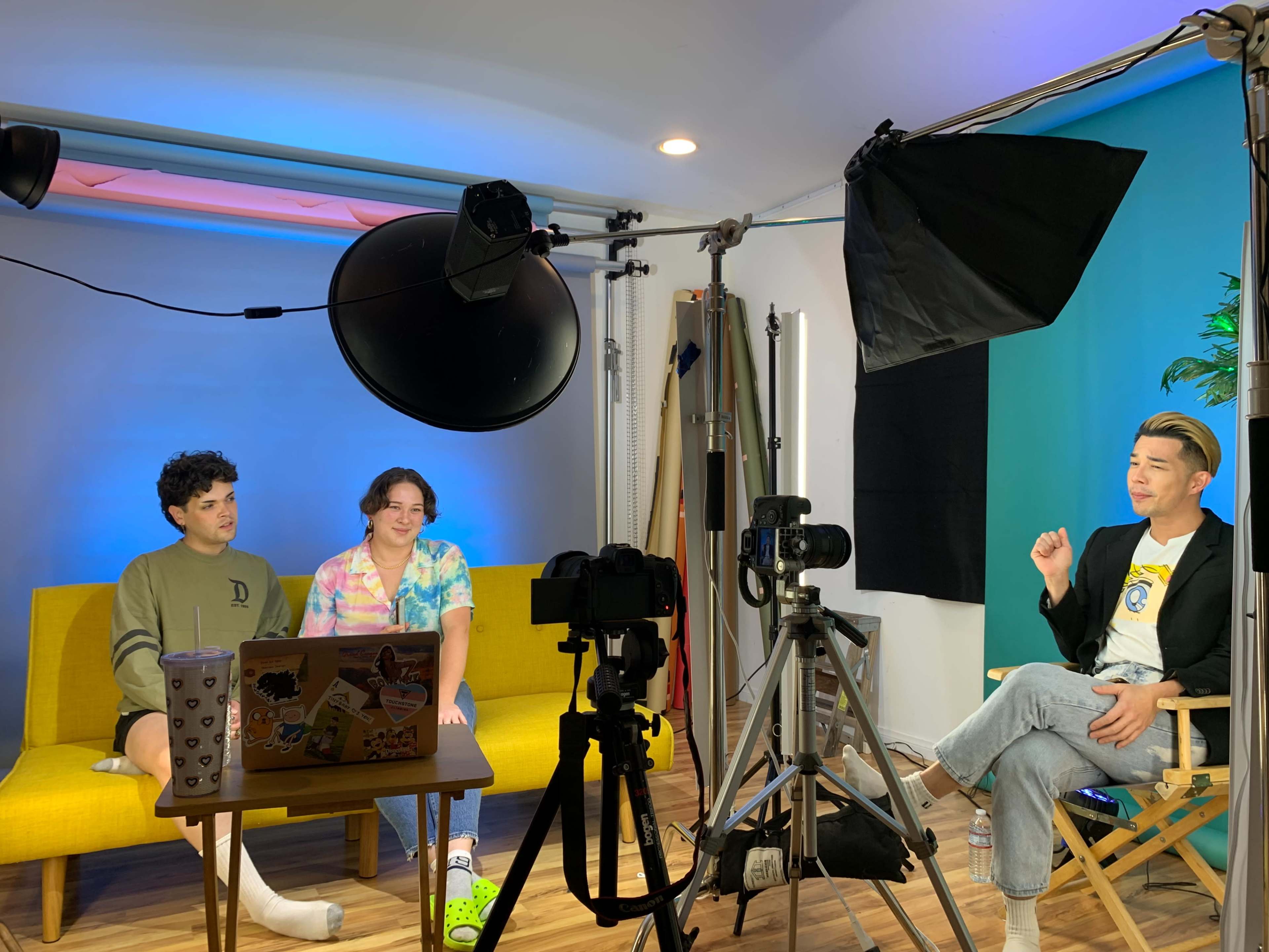 Two individuals are seated on a couch in a brightly lit studio, while a third person gestures from a chair nearby, with cameras and lighting equipment positioned around them.