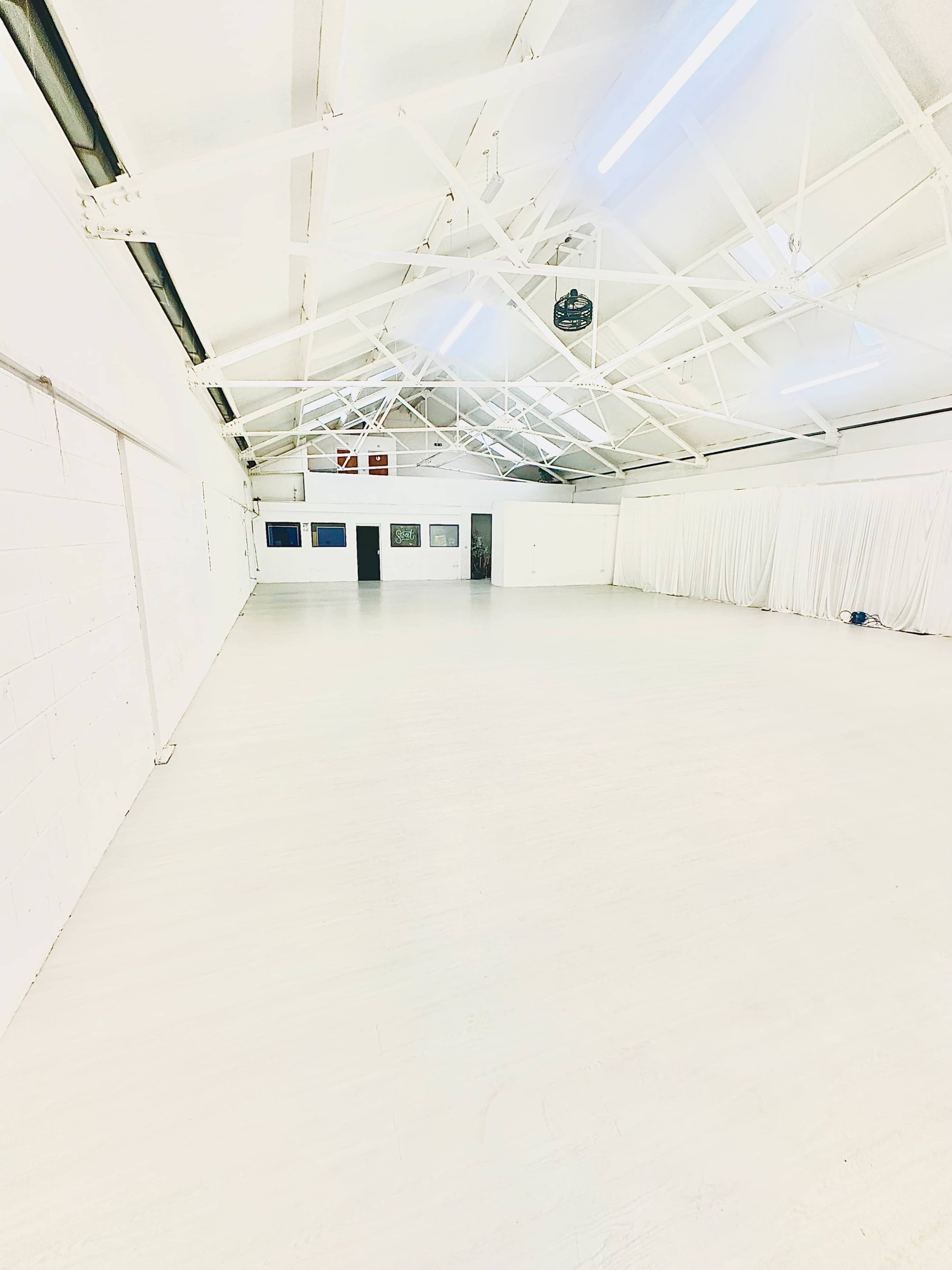 The image shows a large, empty white room with high ceilings and exposed beams, featuring a wooden floor and draped fabric along one wall.