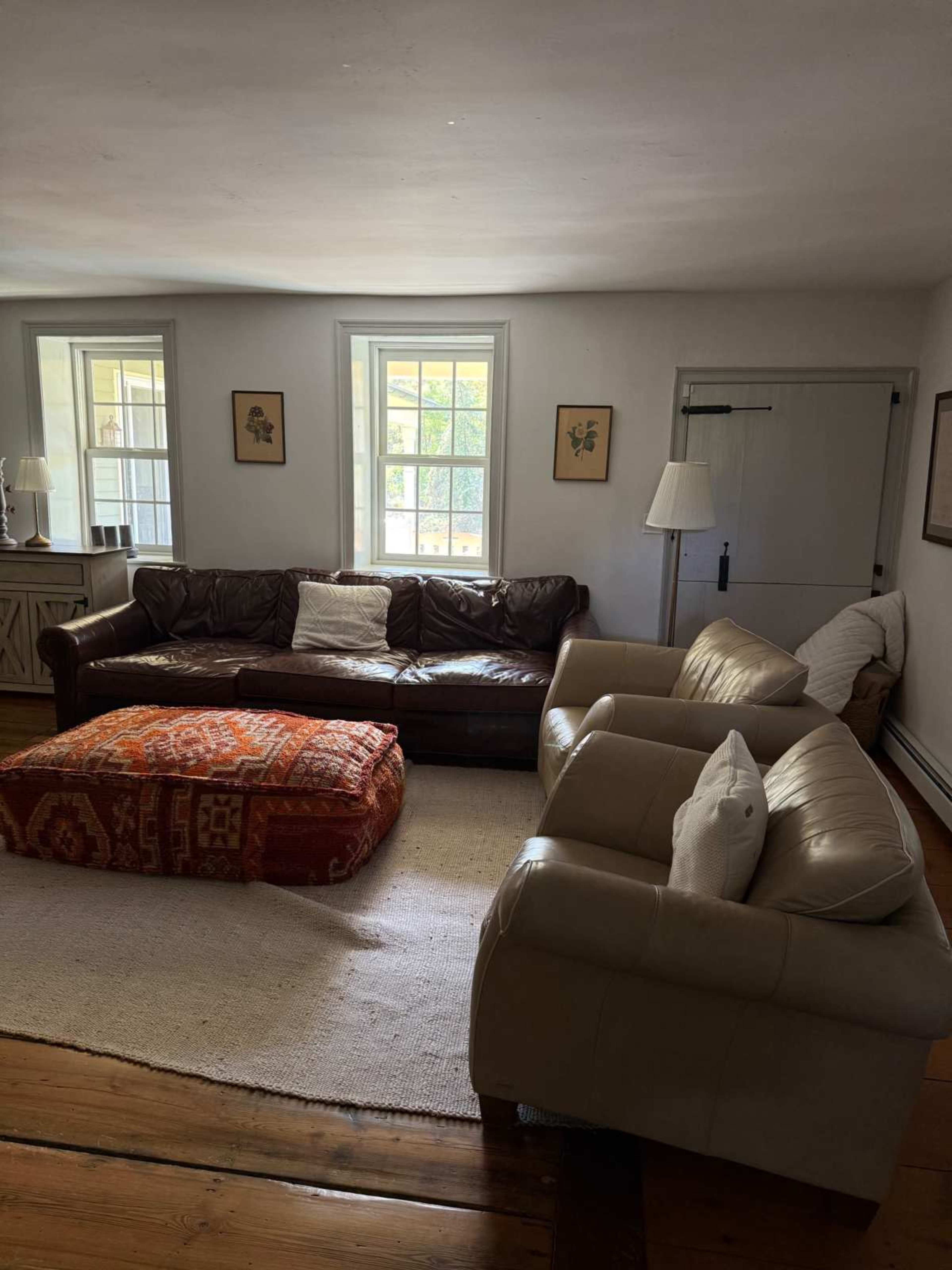 The image shows a cozy living room with a brown leather sofa, two cream-colored armchairs, a patterned ottoman, and a large area rug on a wooden floor.