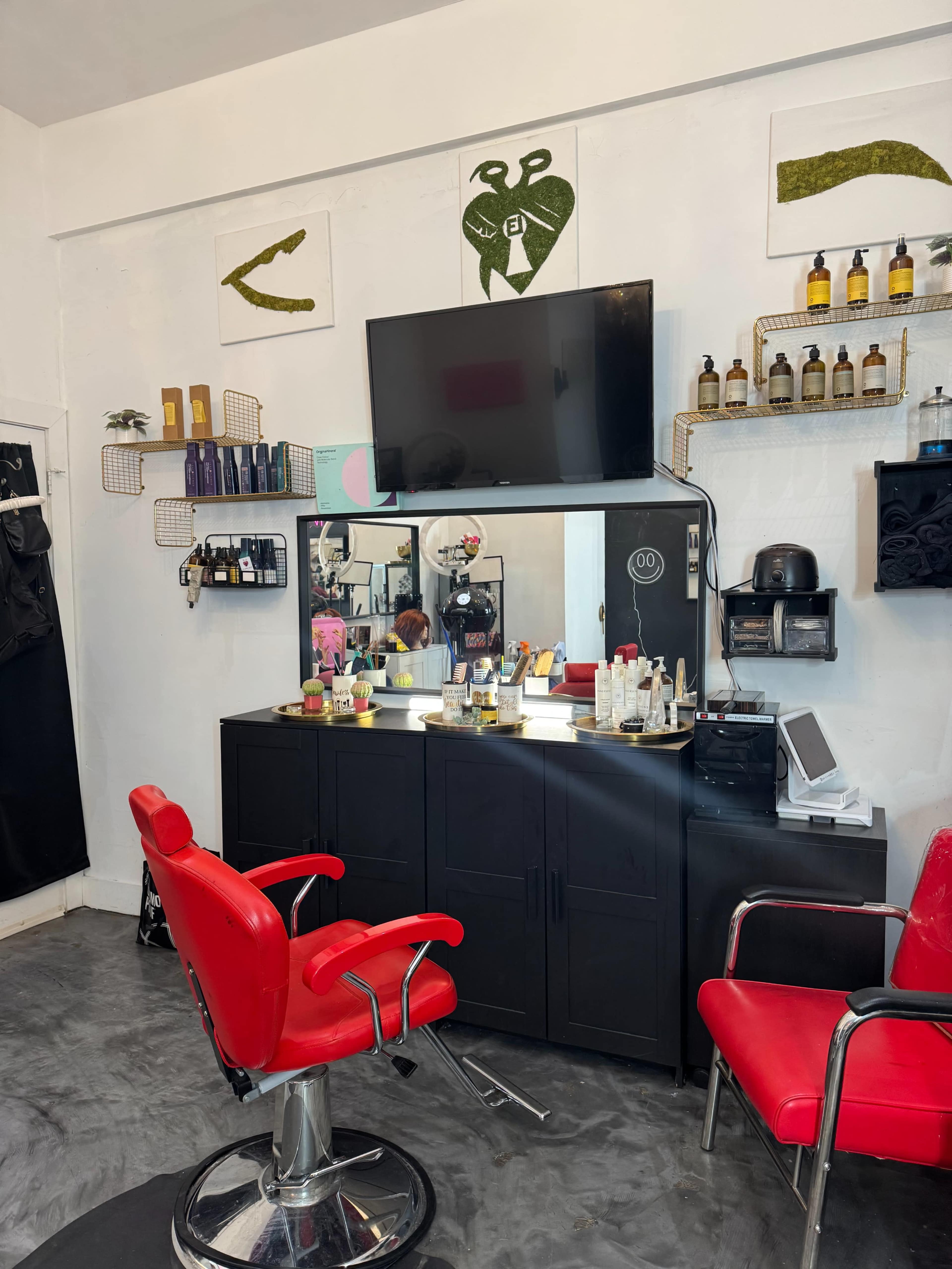 The image shows a tidy salon featuring a black cabinet, a large mirror, a television, and a red salon chair, with various beauty products displayed on the walls and shelves.